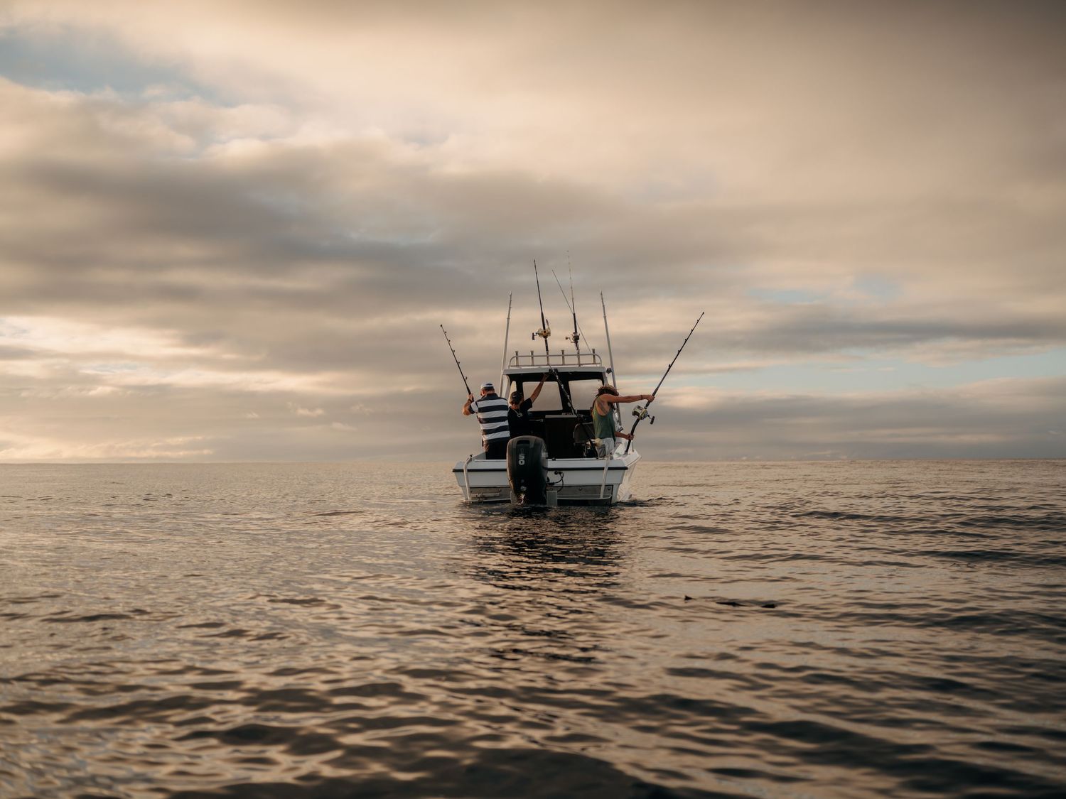 A fishing boat with tall rods sits on calm ocean waters under a dramatic cloudy sky at dusk.