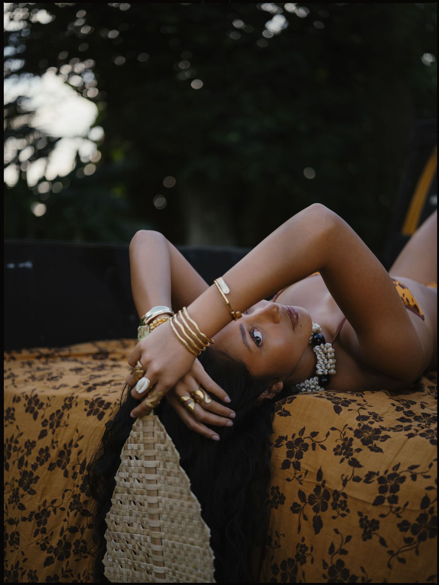 A woman reclines on a floral cushion wearing gold jewelry and bracelets, posing elegantly outdoors.