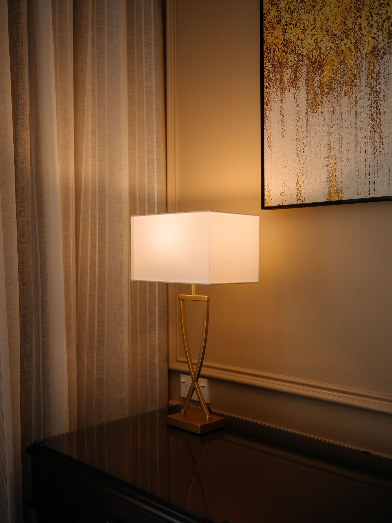 Elegant gold table lamp with white square shade on dark dresser beside sheer curtains and abstract wall art.