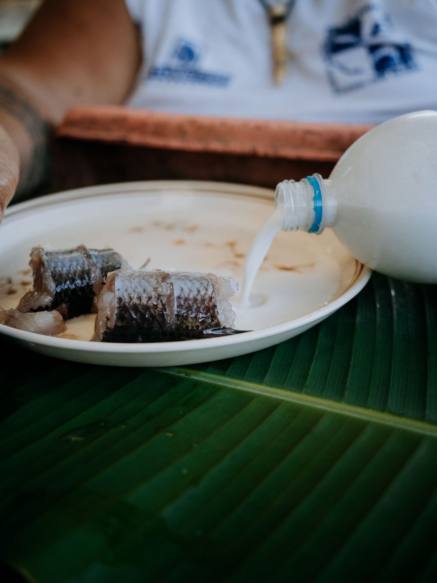 White milk being poured over grilled fish pieces on a plate resting on a banana leaf.