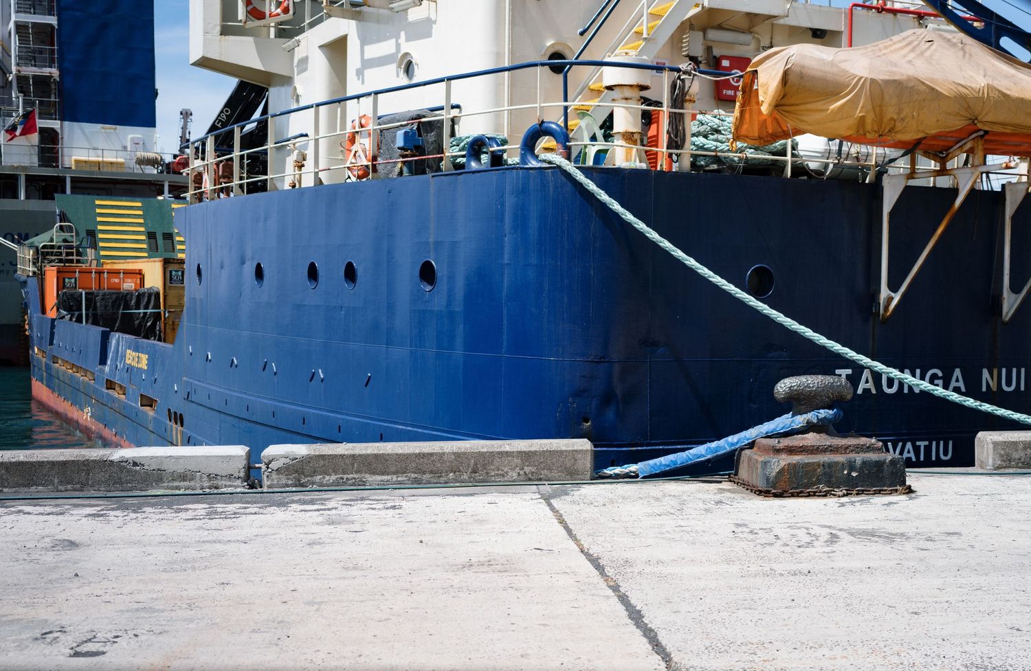 Blue cargo ship Taunga Nui docked at port, secured with mooring lines on a clear day.