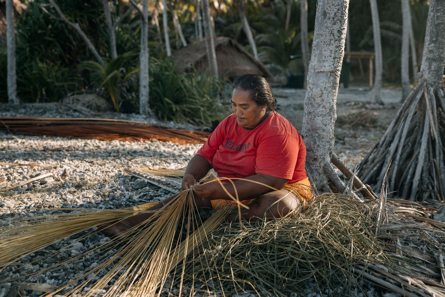 Woman in red shirt weaving dried grass bundles outdoors near palm trees on a tropical beach.