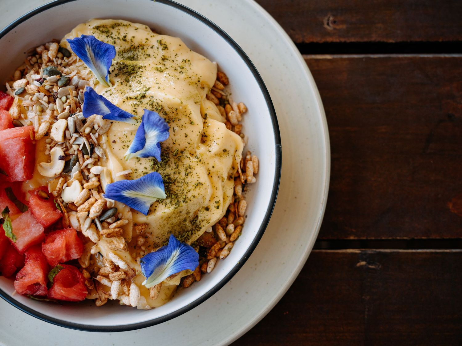 Overhead view of a healthy smoothie bowl with watermelon, banana, granola, and blue edible flowers on a wooden table.