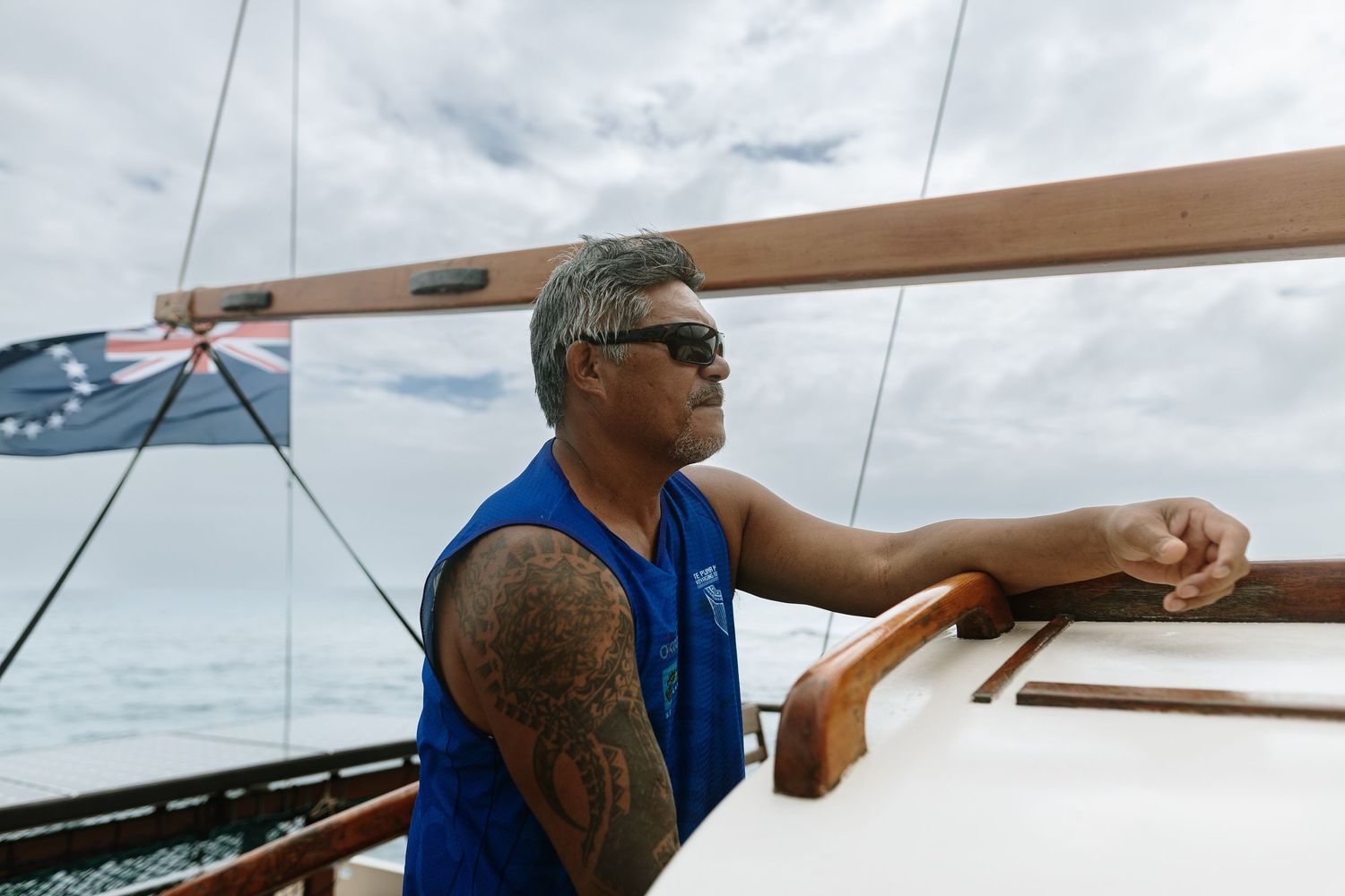 Tattooed Polynesian man steering a wooden sailboat on the ocean, wearing a blue vest and sunglasses.