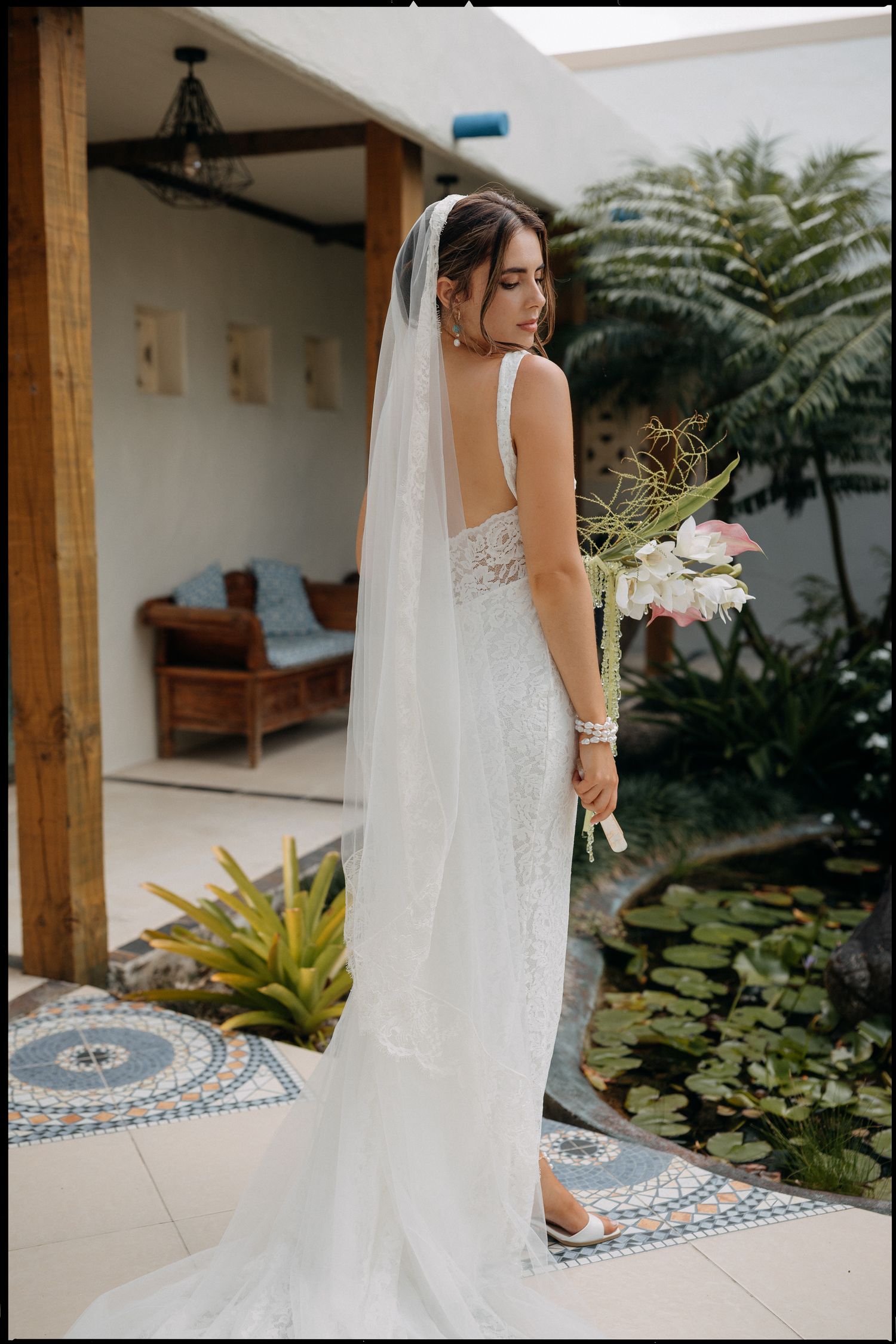Bride in backless lace wedding gown with long veil holding bouquet, standing in tropical garden courtyard.