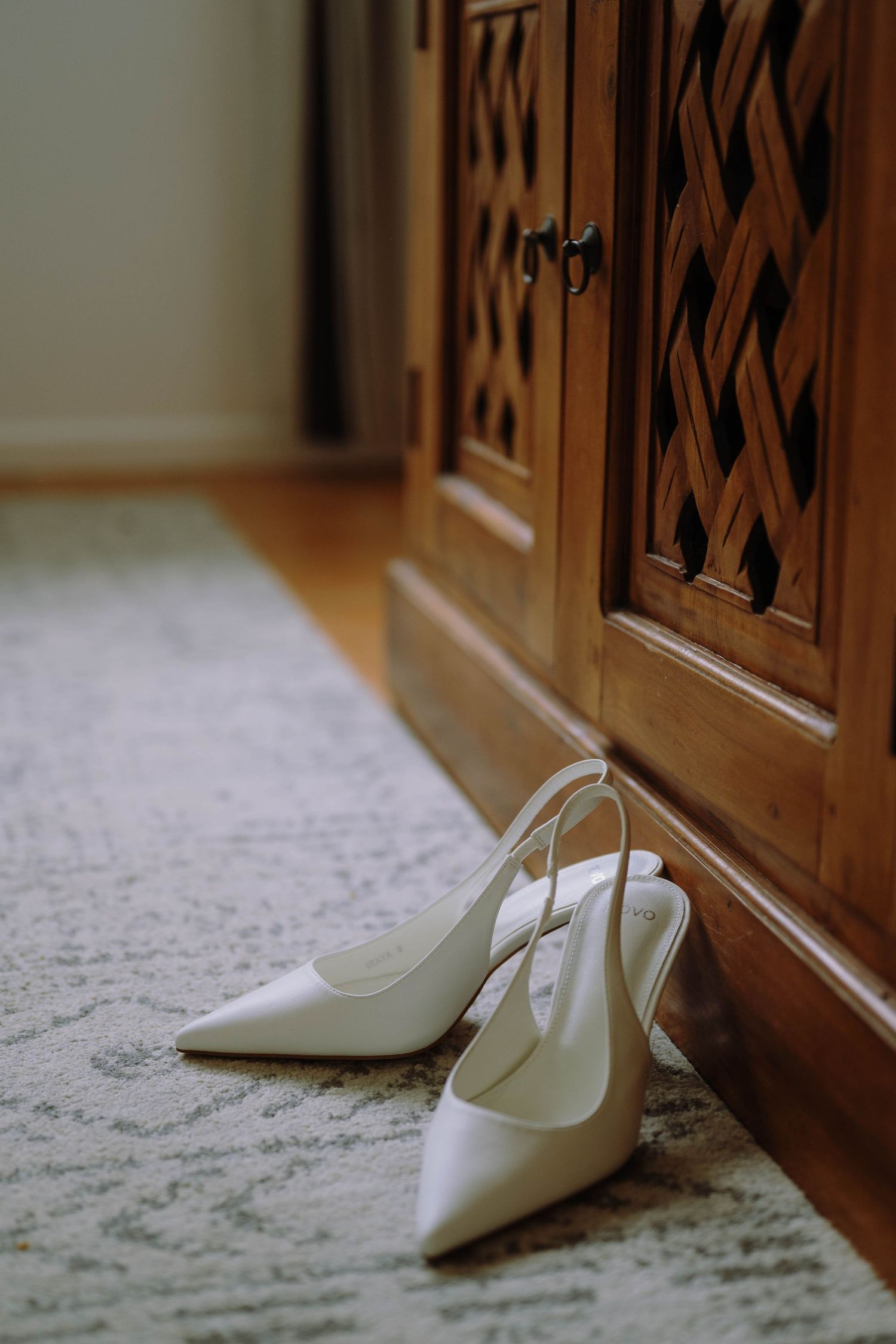 Elegant white slingback heels placed on a rug beside a ornate wooden cabinet, perfect bridal wedding shoes.