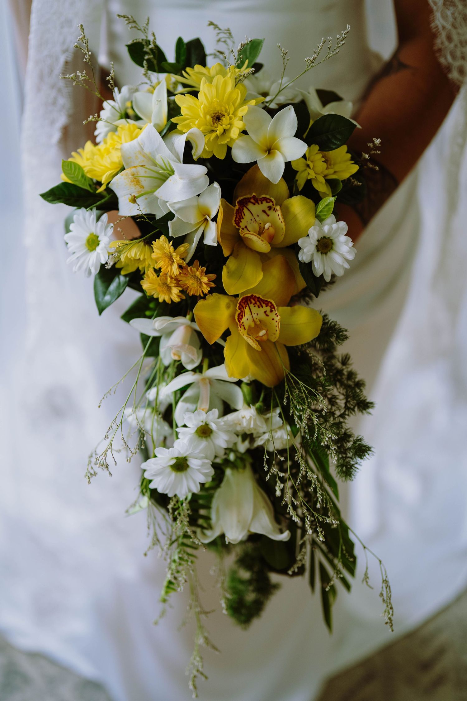 Bride holding a cascading wedding bouquet with yellow orchids, white lilies, daisies, and greenery in an elegant arrangement.