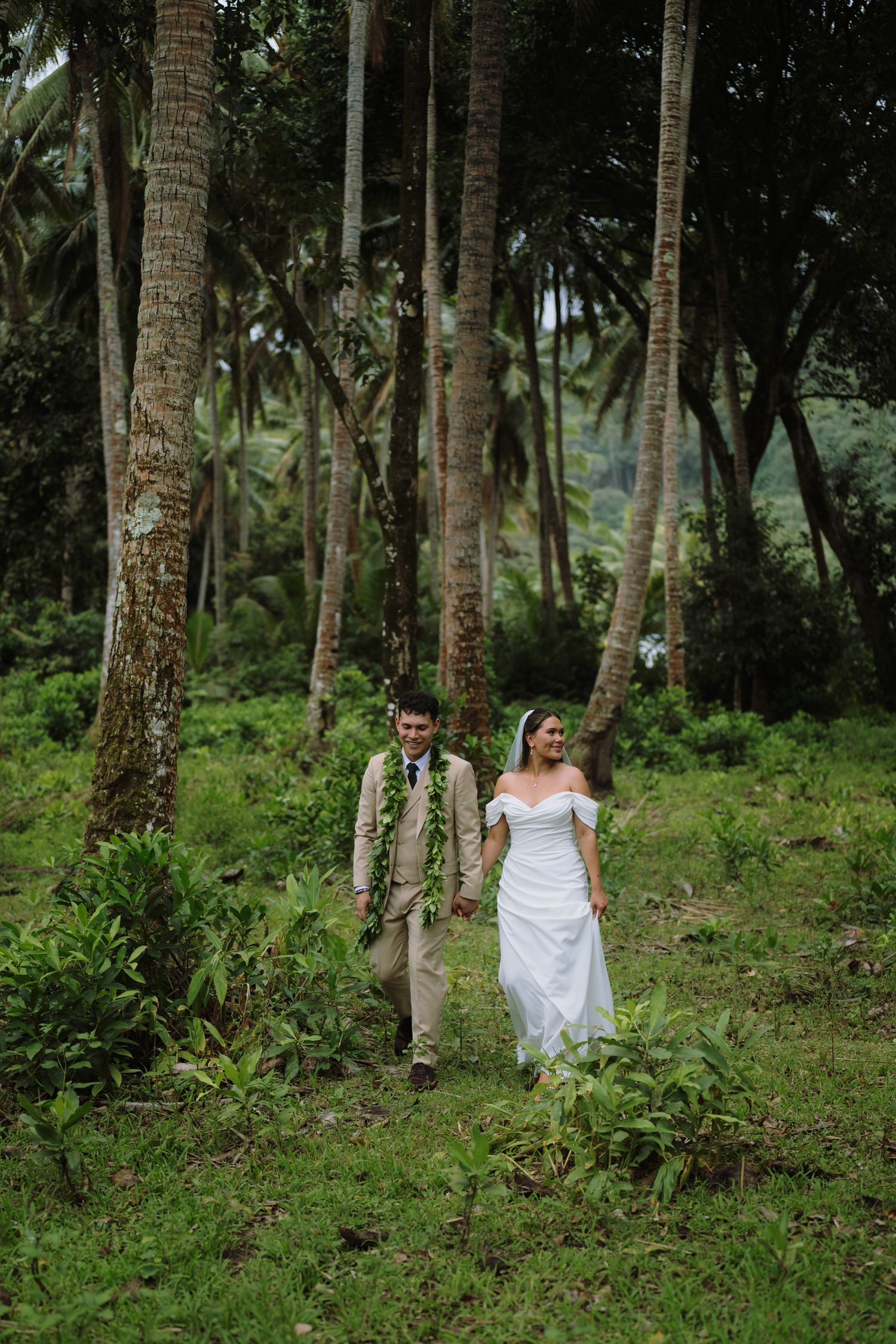 Bride and groom walk hand-in-hand through a lush tropical palm forest in wedding attire.