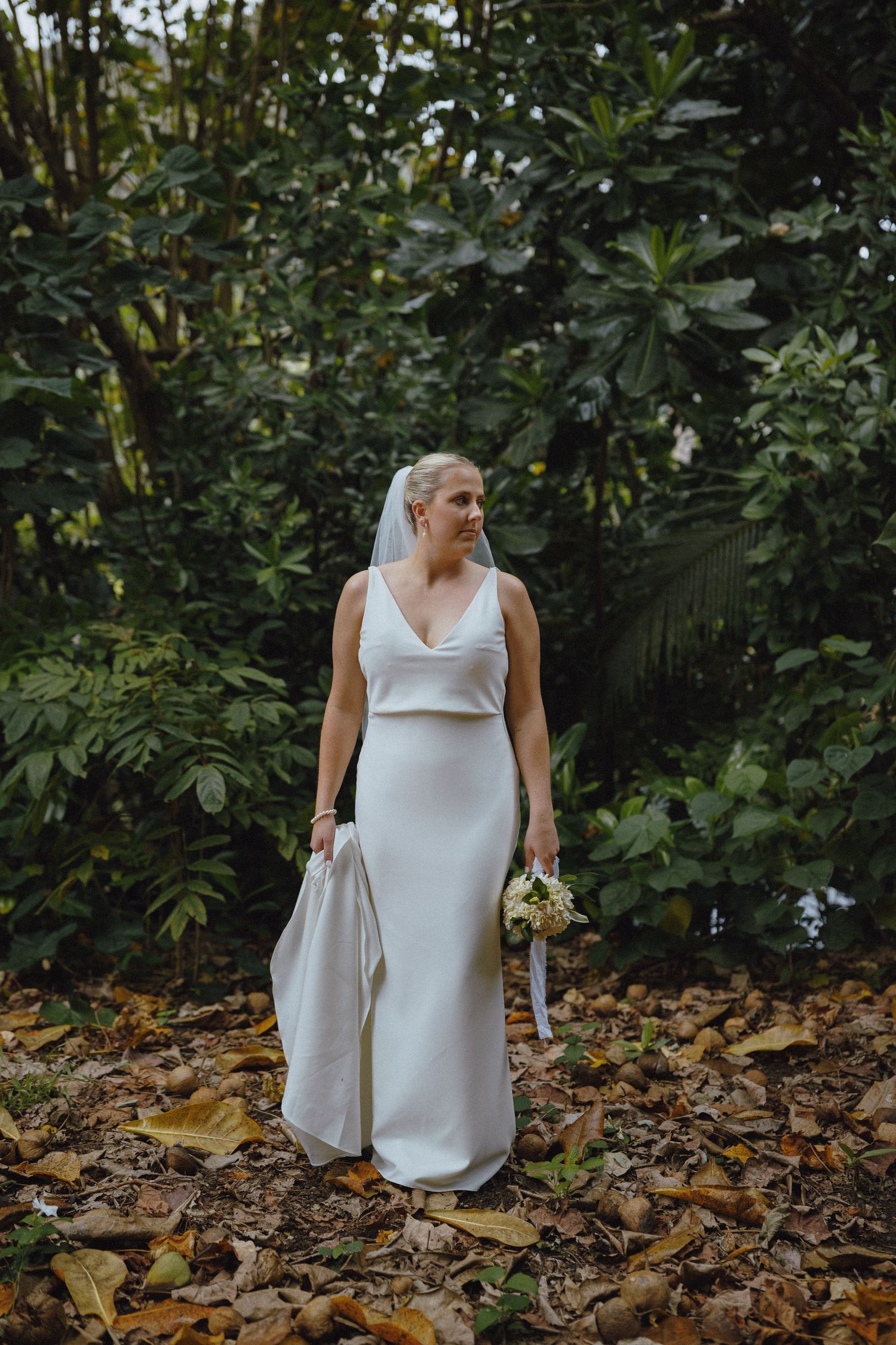 Bride in white gown and veil holds bouquet among lush tropical greenery on fallen autumn leaves.