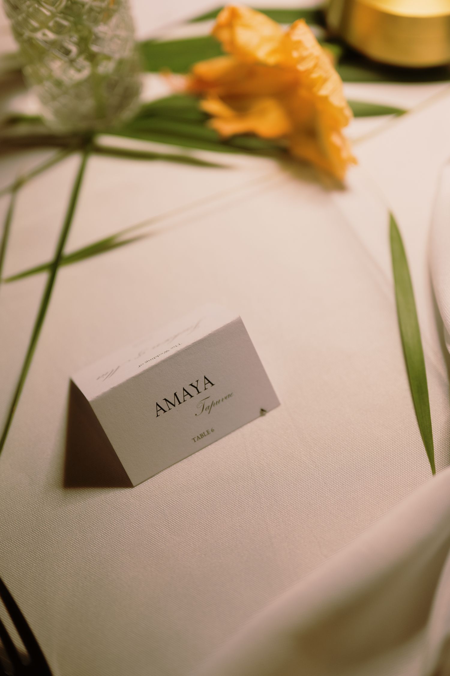 Elegant place card reading AMAYA on a white linen tablecloth beside a yellow iris flower and green leaves.