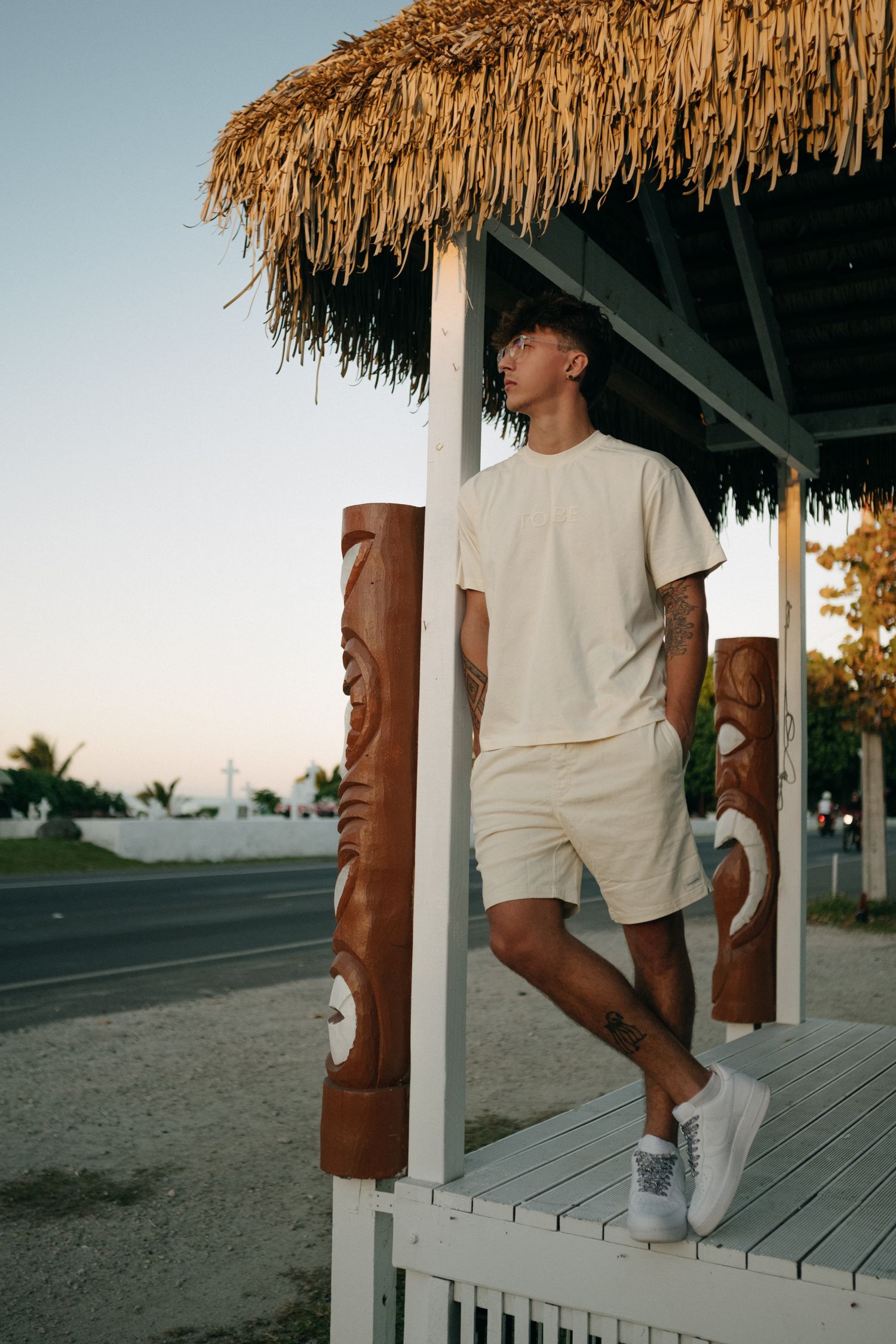 Young man in beige outfit standing under tiki hut with carved wooden posts near roadside at dusk.