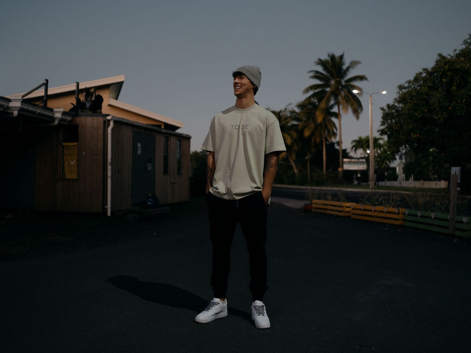 Young man in beanie and white sneakers stands on dark pavement at dusk near palm trees and buildings.