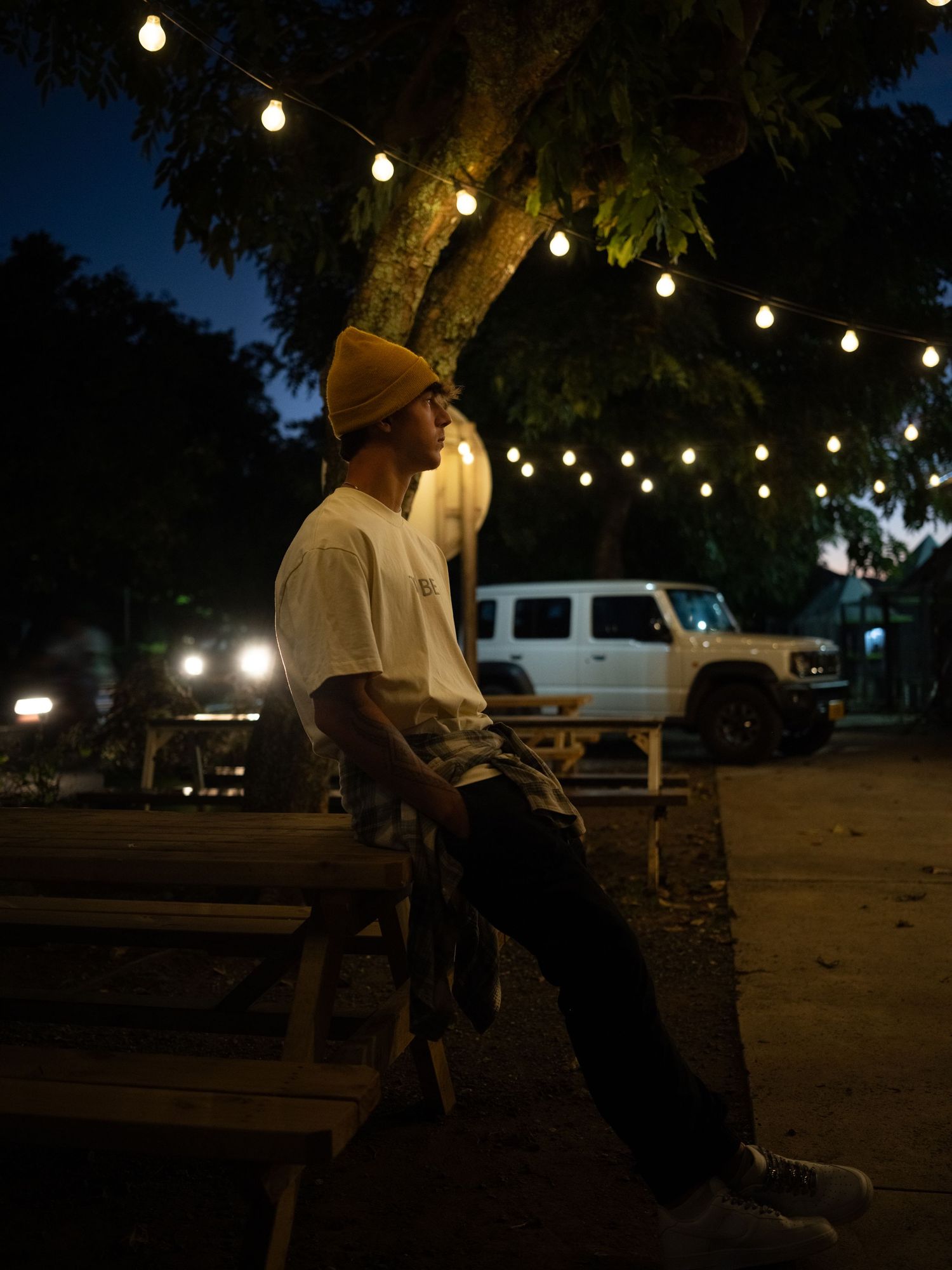 Man in yellow beanie sits on bench under string lights at night with white SUV in background.