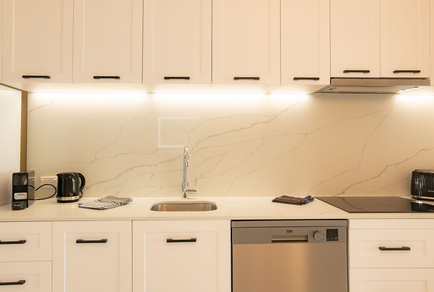 Modern white kitchen with marble backsplash, under-cabinet lighting, and stainless steel dishwasher.