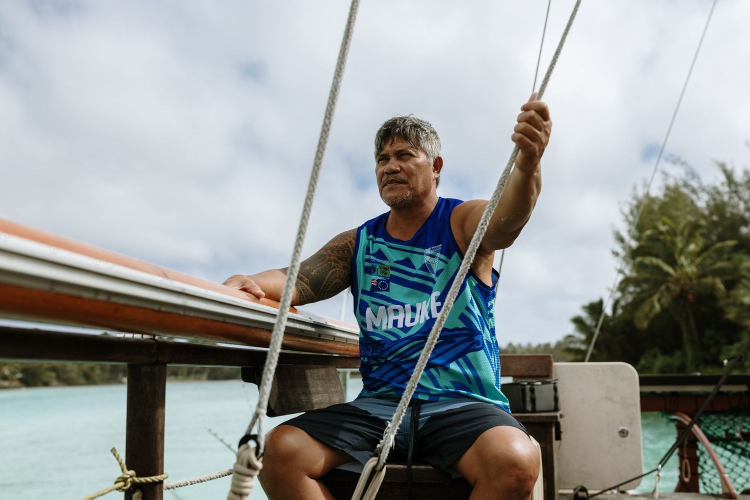 Man in blue tank top sailing a boat in tropical waters, holding rigging ropes on a sunny cloudy day.