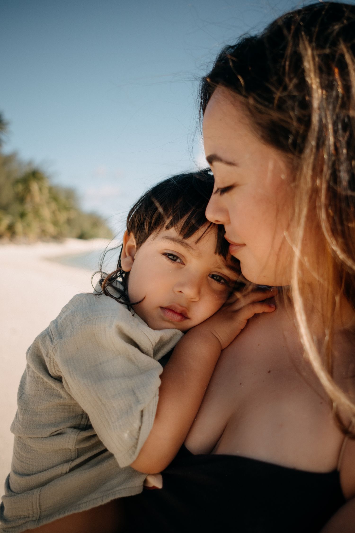 A mother tenderly holds her young child on a sunny beach, sharing a gentle moment with palm trees in the background.