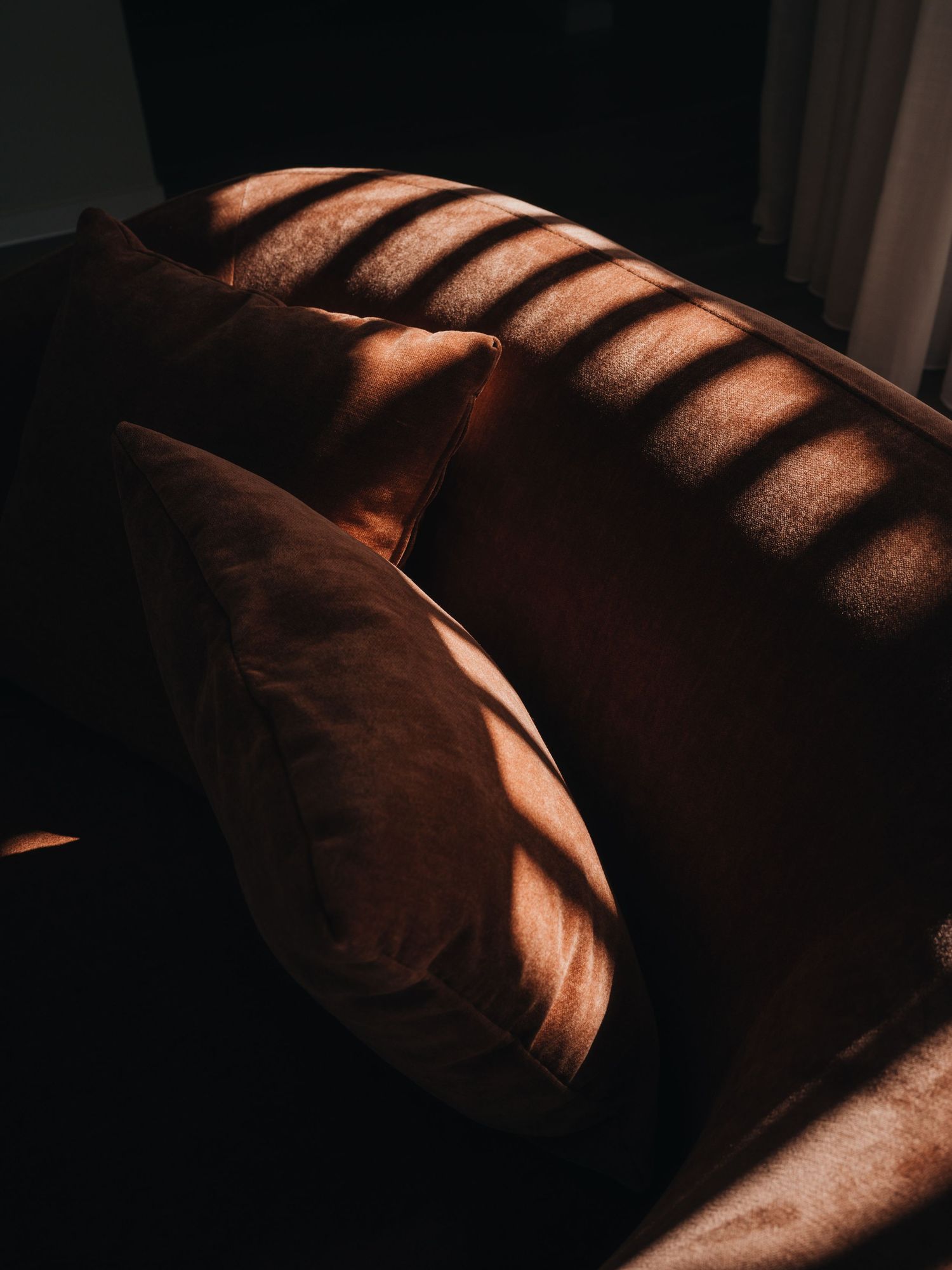 Warm brown velvet sofa cushions bathed in dramatic striped shadows from sunlight filtering through blinds.