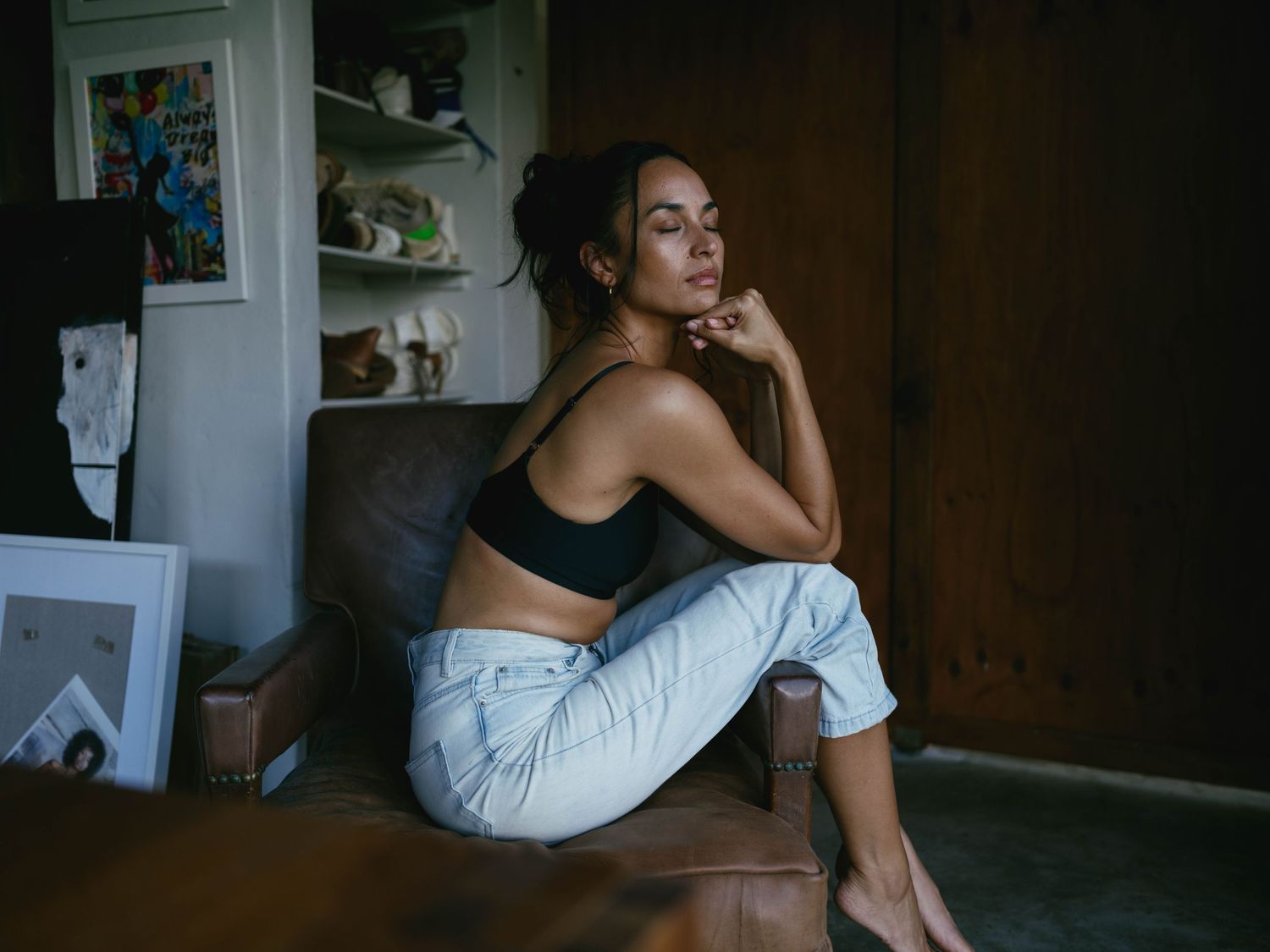 A young woman in a black crop top and light jeans sits thoughtfully on a chair in a cozy, dimly lit room.