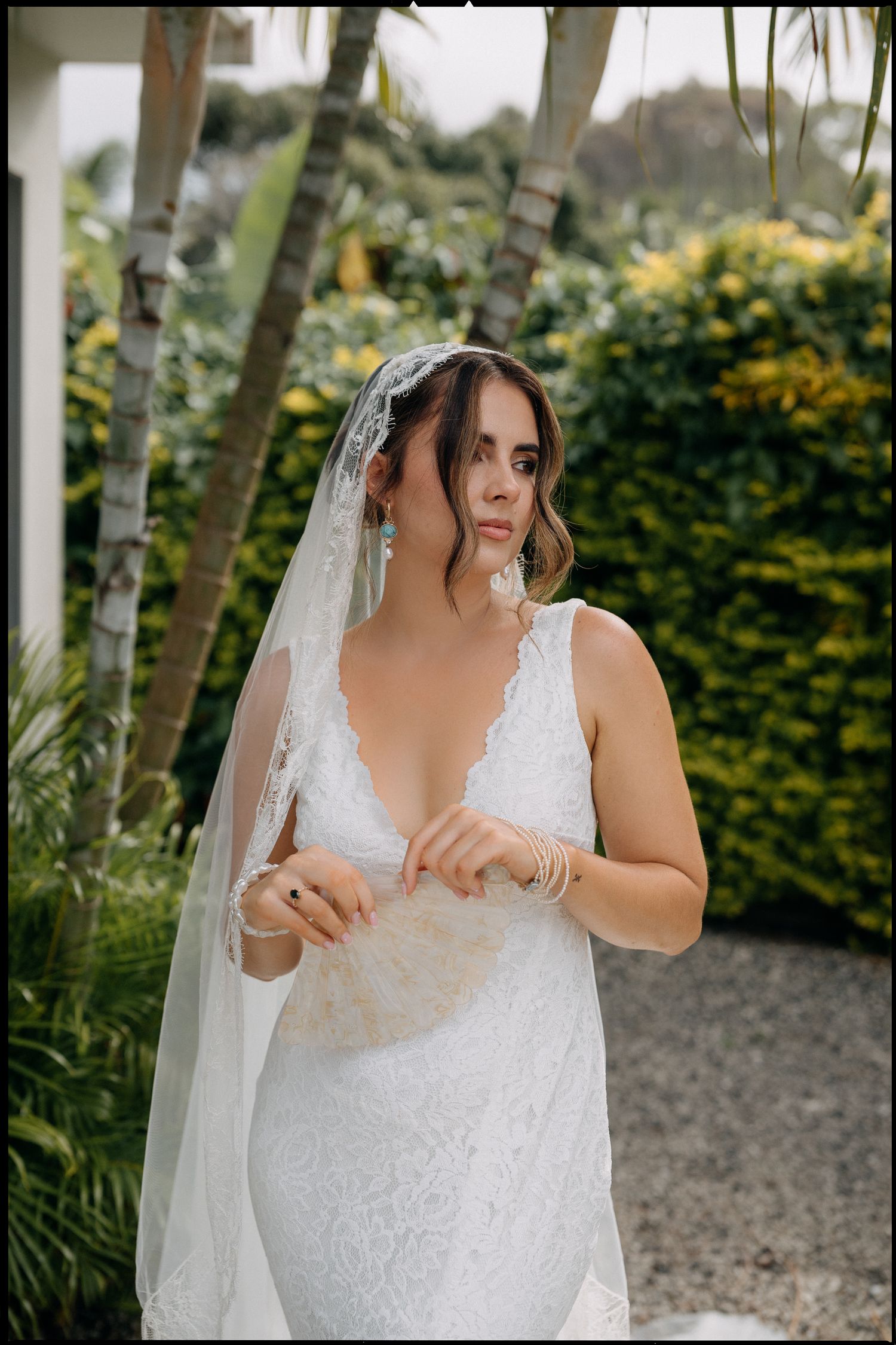 Bride in lace wedding dress and veil holds fan bouquet outdoors among tropical palms and greenery.