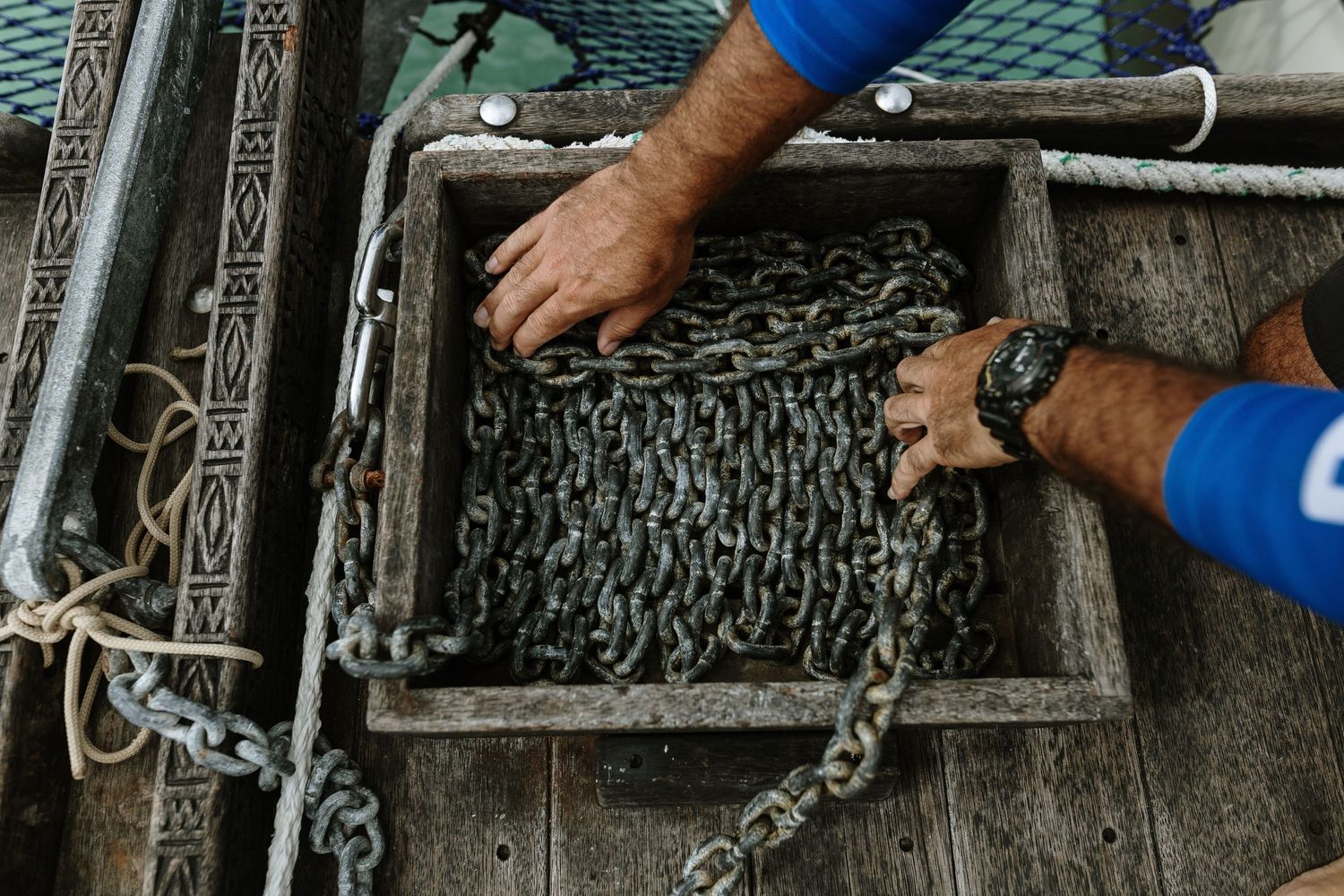 Two hands arranging rusty metal chains inside a weathered wooden box on a boat deck.