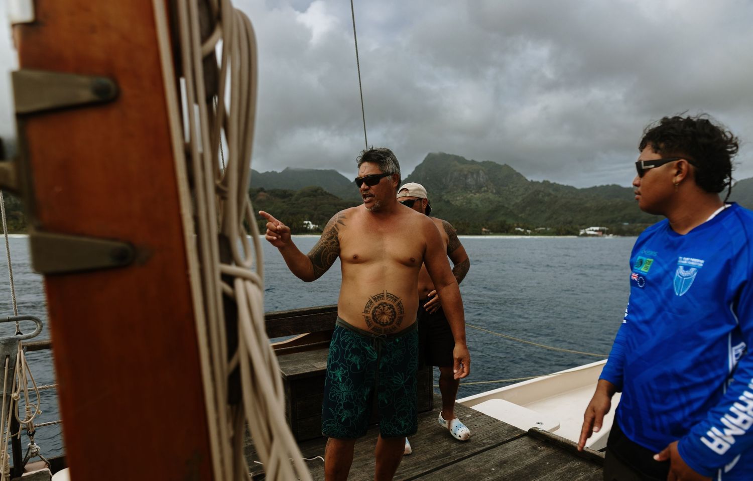 Three shirtless men on a wooden boat deck with tropical green hills in the background, one pointing into the distance.