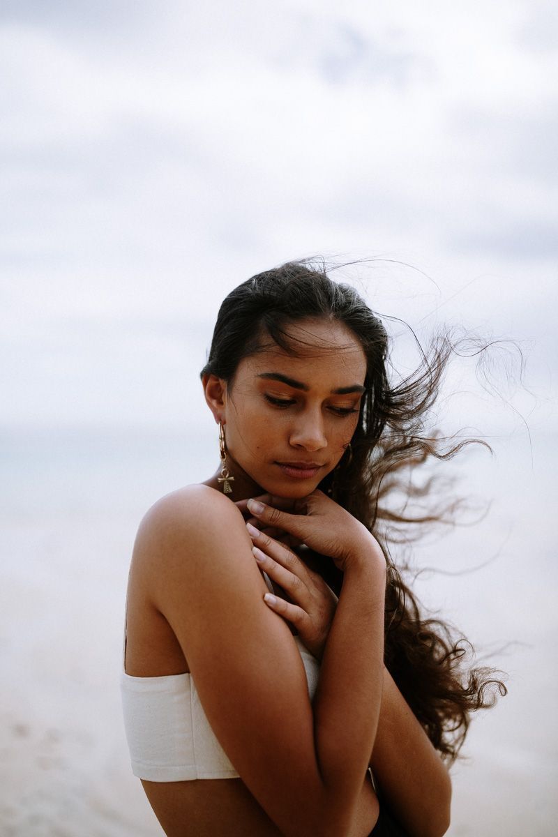Young woman in white bandeau top at beach, wind blowing long dark hair, eyes closed, hands crossed over chest.