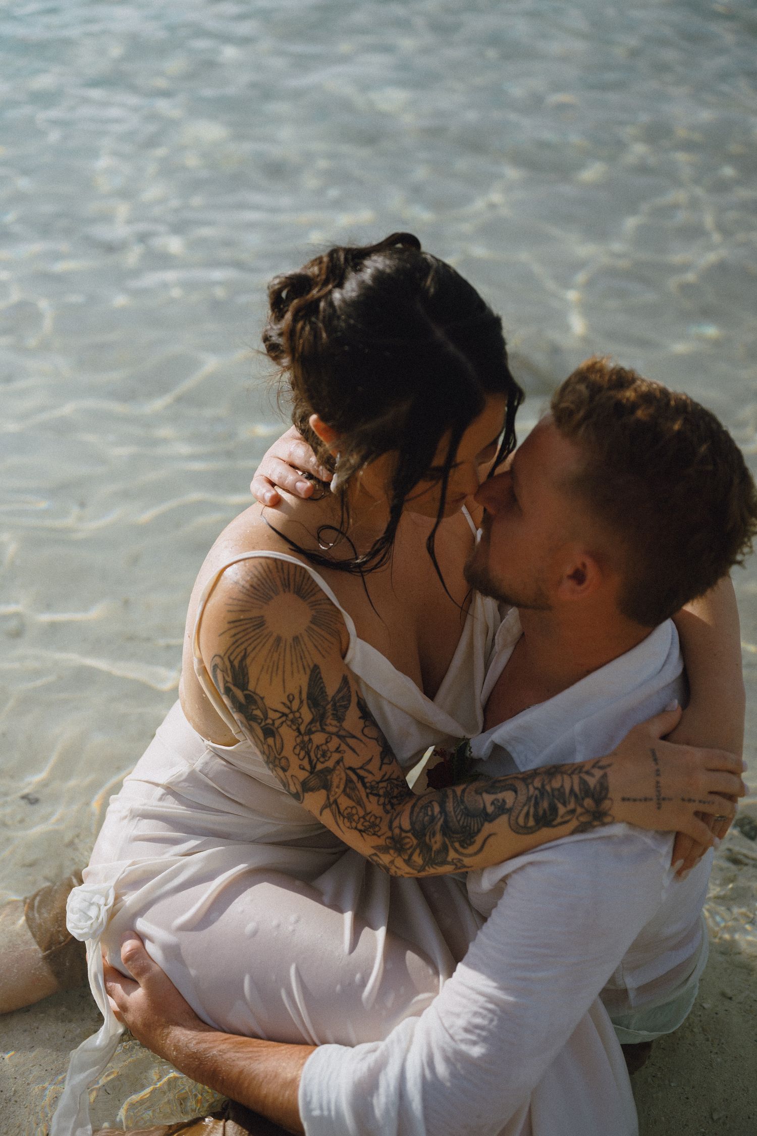 Couple sharing a romantic kiss on a sandy beach shore, woman with floral tattoos in white dress embracing tattooed man.