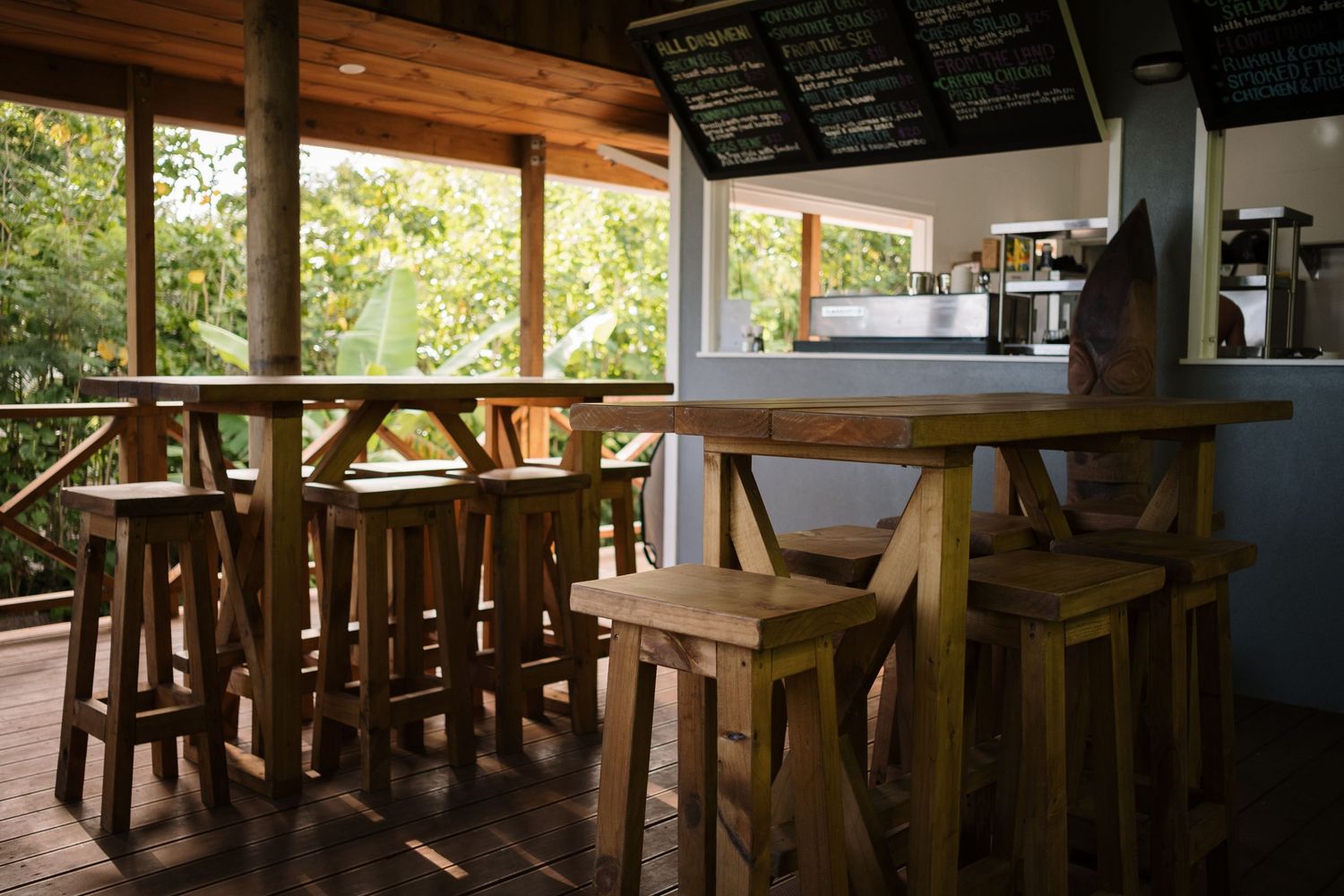 Rustic wooden bar stools and high tables in an open-air tropical cafe with lush green views.
