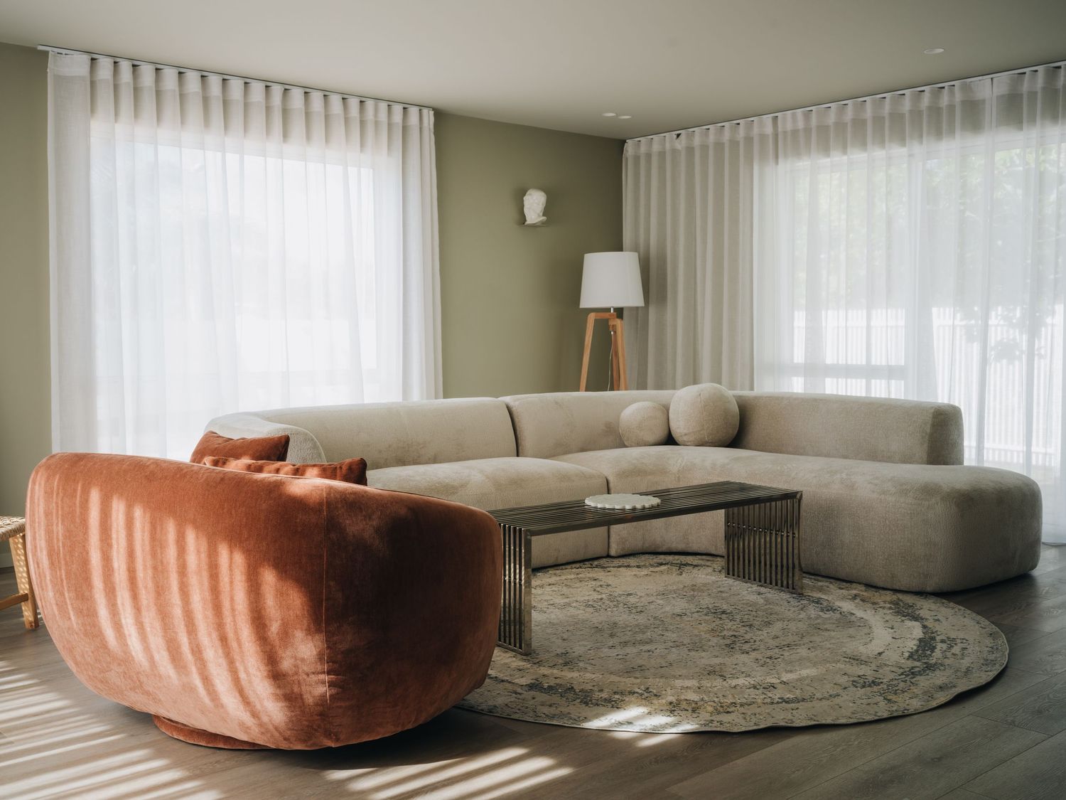 Modern living room with beige curved sectional sofa, terracotta armchair, round rug, and sheer white curtains.