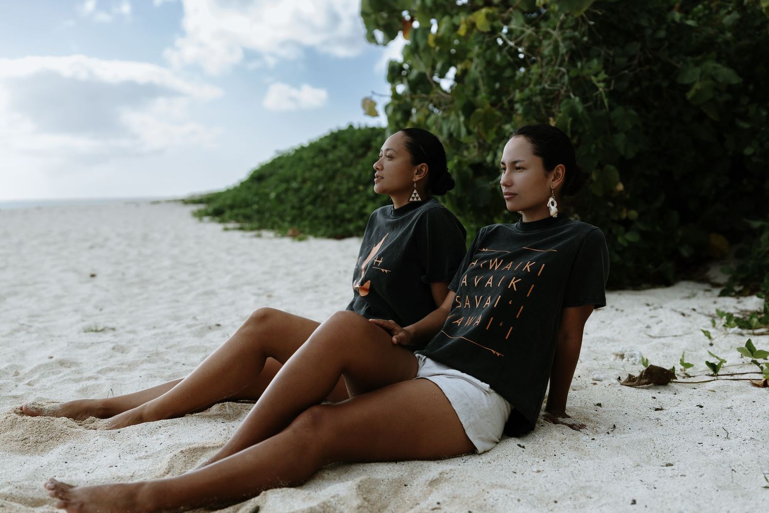 Two women in black shirts sit relaxed on white sandy beach with lush green trees behind them.