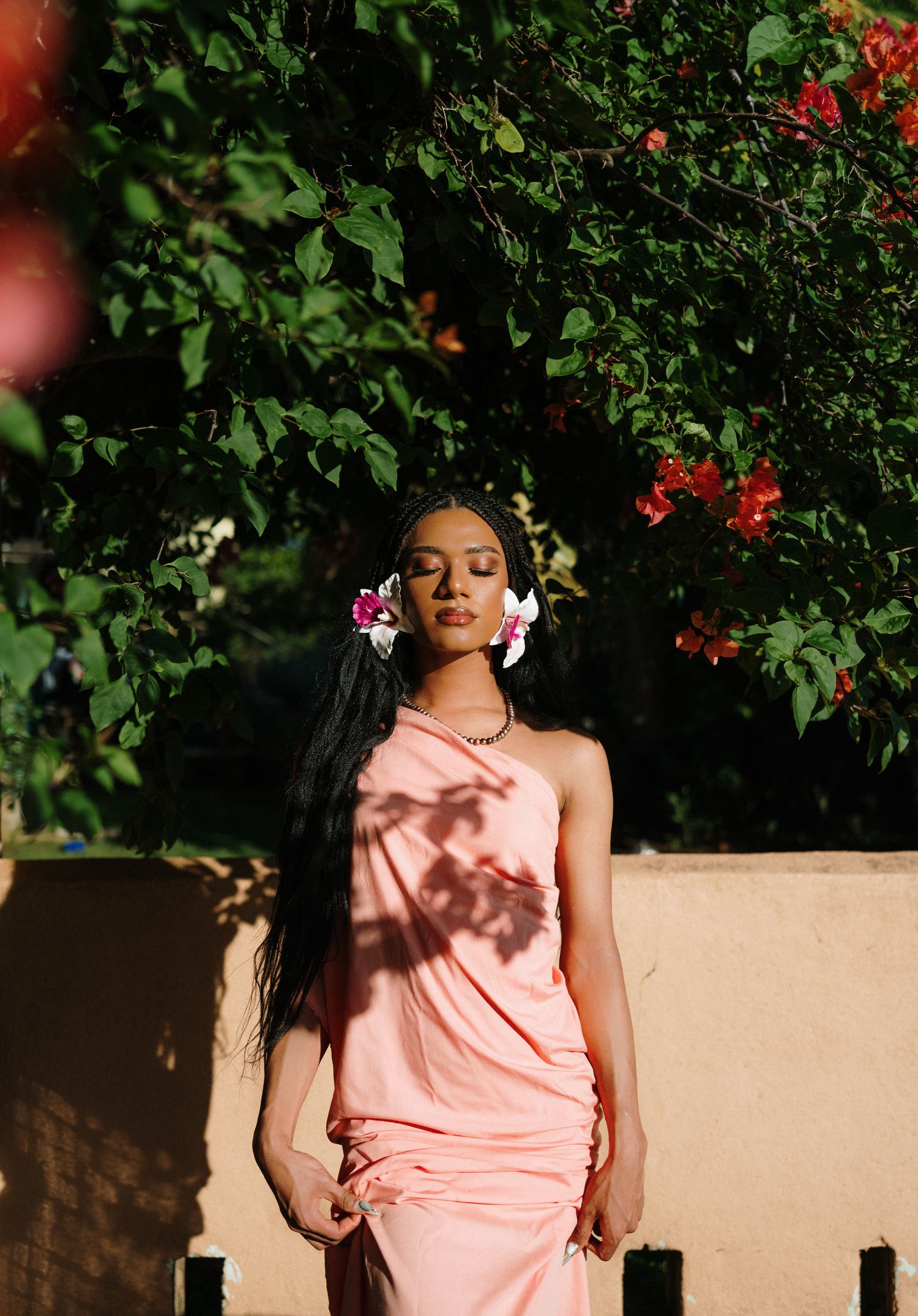 Woman in peach one-shoulder dress with floral hair accessories poses outdoors near lush green and red flowering trees.