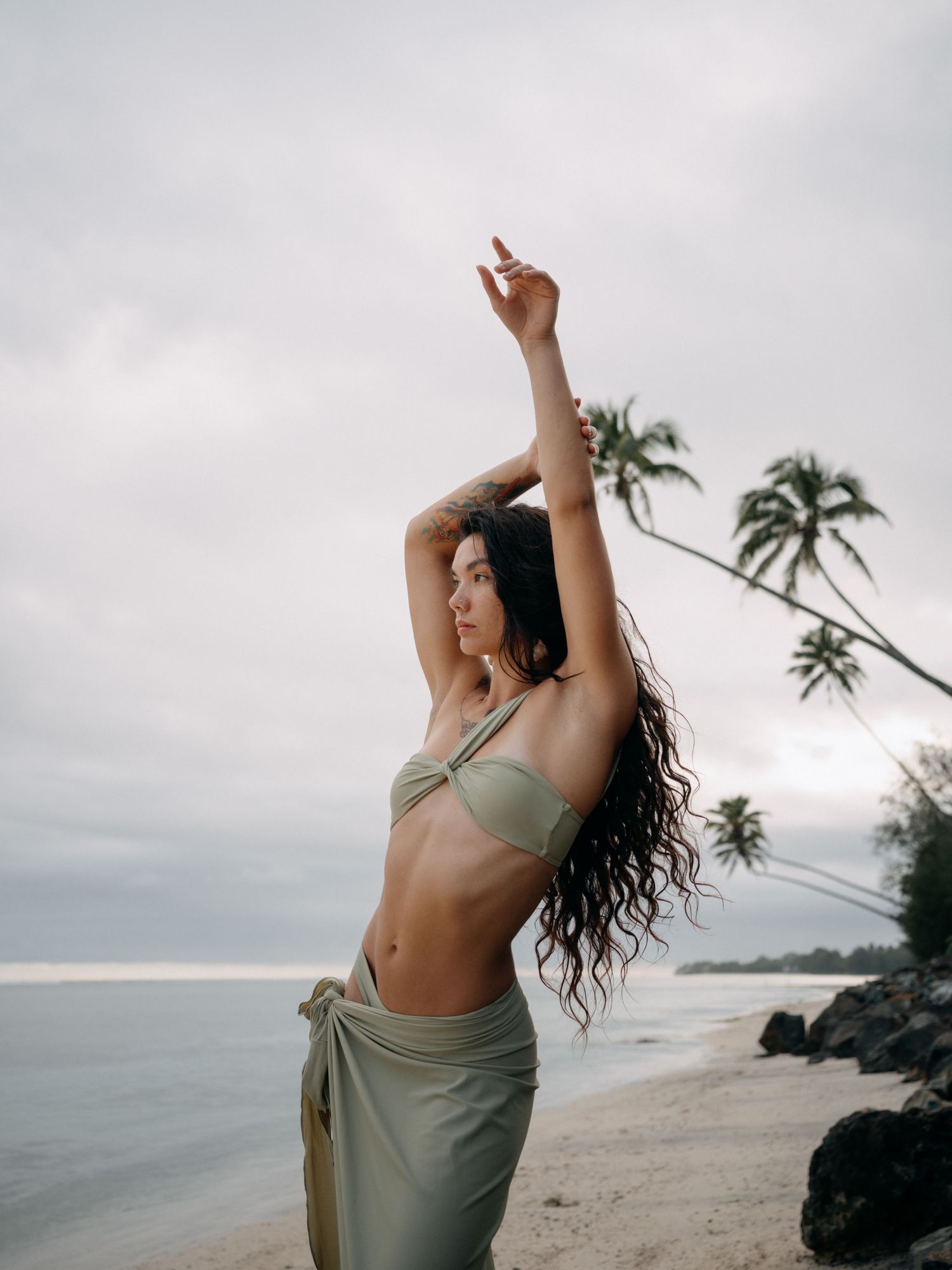 Woman in olive green bikini top and sarong poses on tropical beach with palm trees and overcast sky.