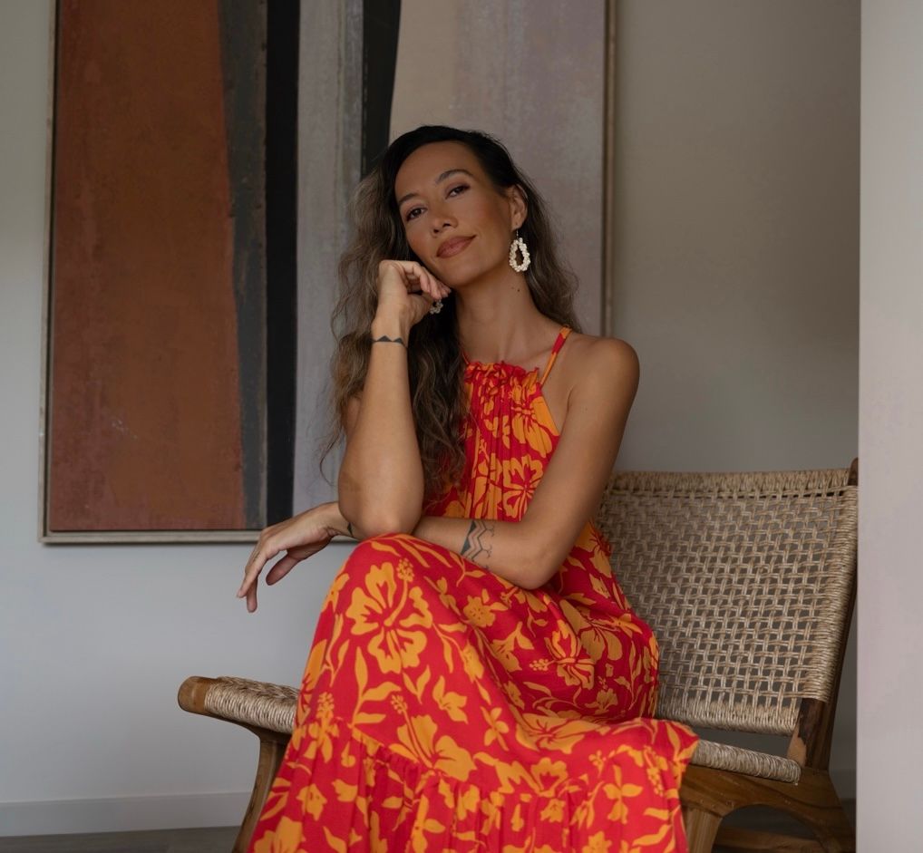 Woman in red floral maxi dress sitting on rattan chair, wearing gold hoop earrings, posing elegantly indoors.