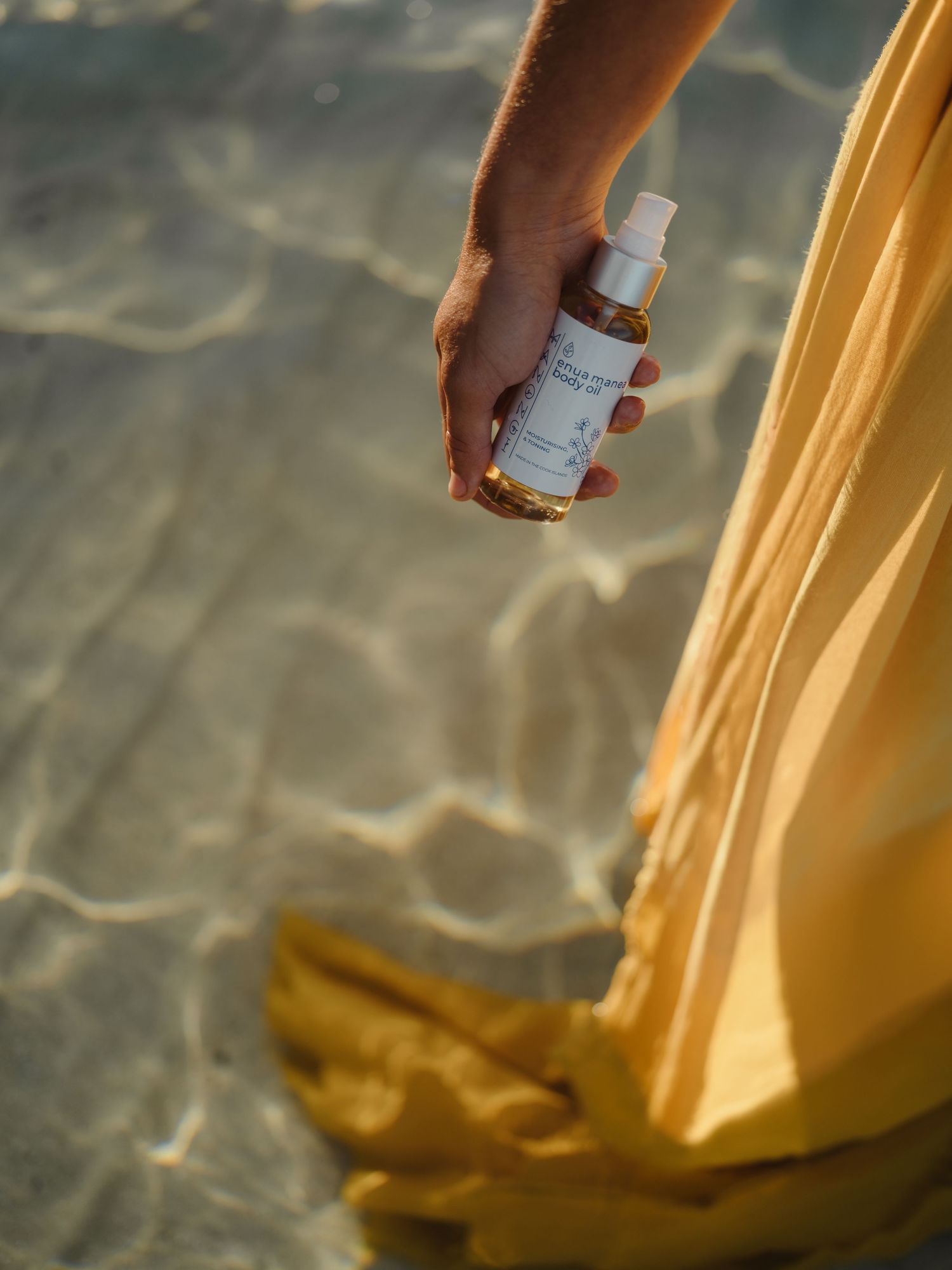 A hand holds a small spray bottle near shimmering shallow water, beside a flowing yellow dress in golden sunlight.