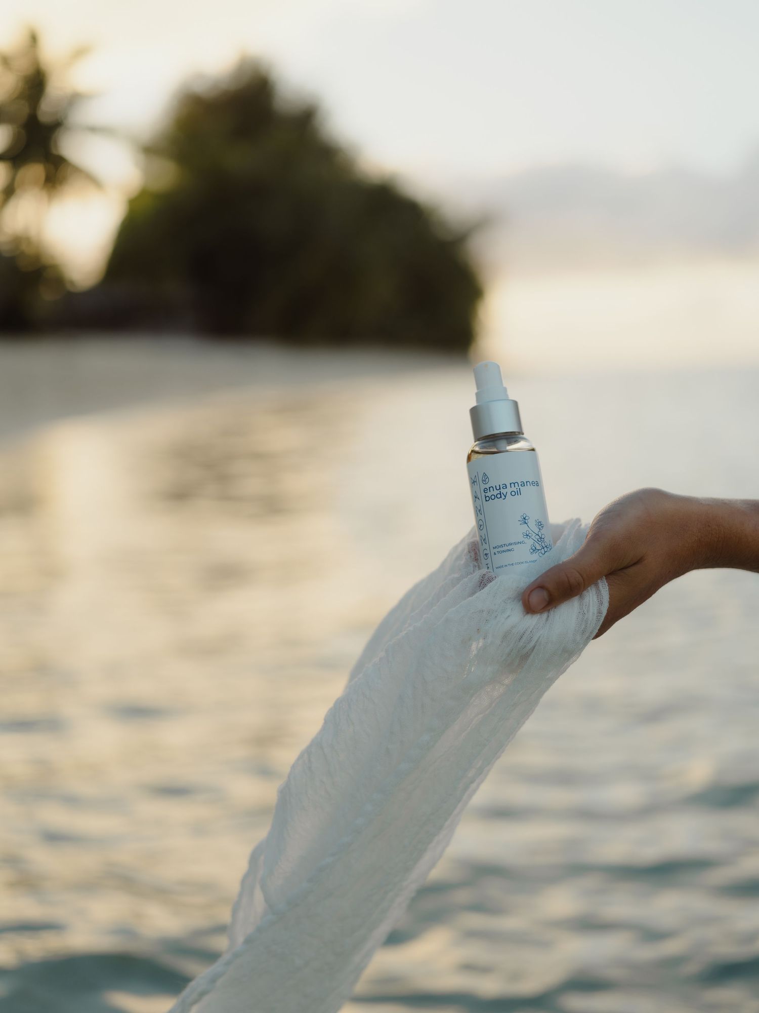 Hand holding a skincare serum bottle wrapped in white fabric against a serene tropical beach sunset backdrop.