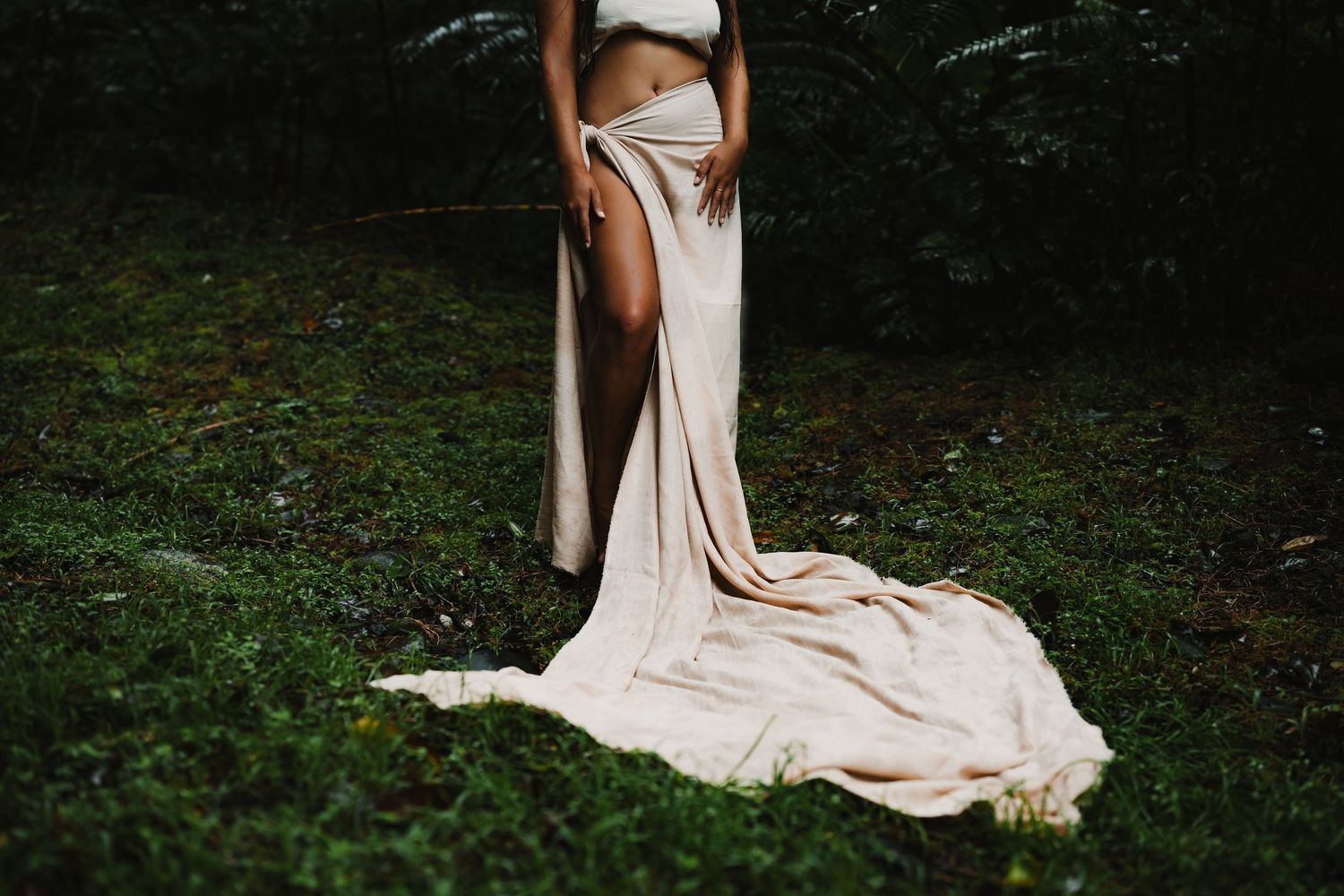 Pregnant woman in white crop top and flowing beige maxi skirt with high slit, standing outdoors on green grass.