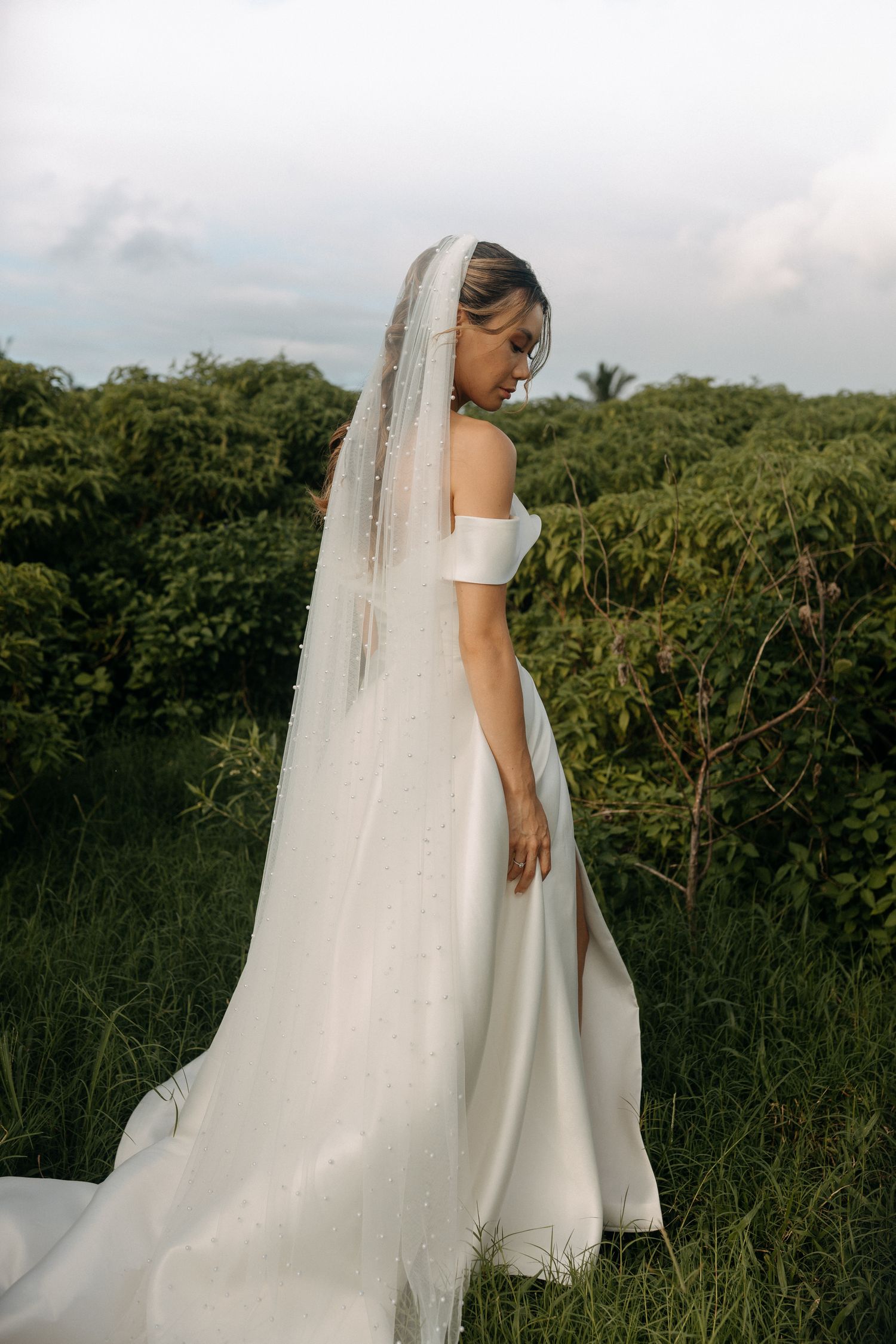 Bride in elegant off-shoulder white gown with pearl-adorned veil, standing in lush green garden setting.