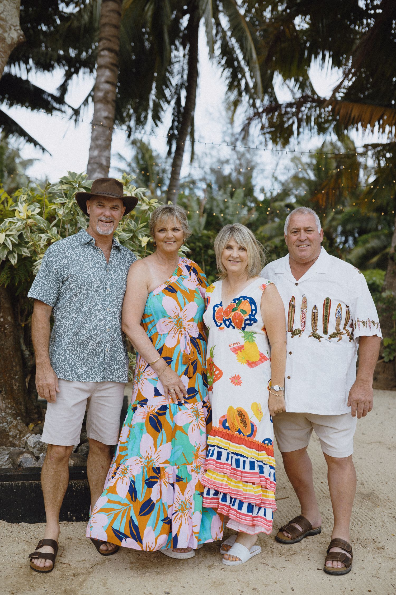 Two couples posing together on a tropical beach surrounded by palm trees, wearing colorful summer outfits.