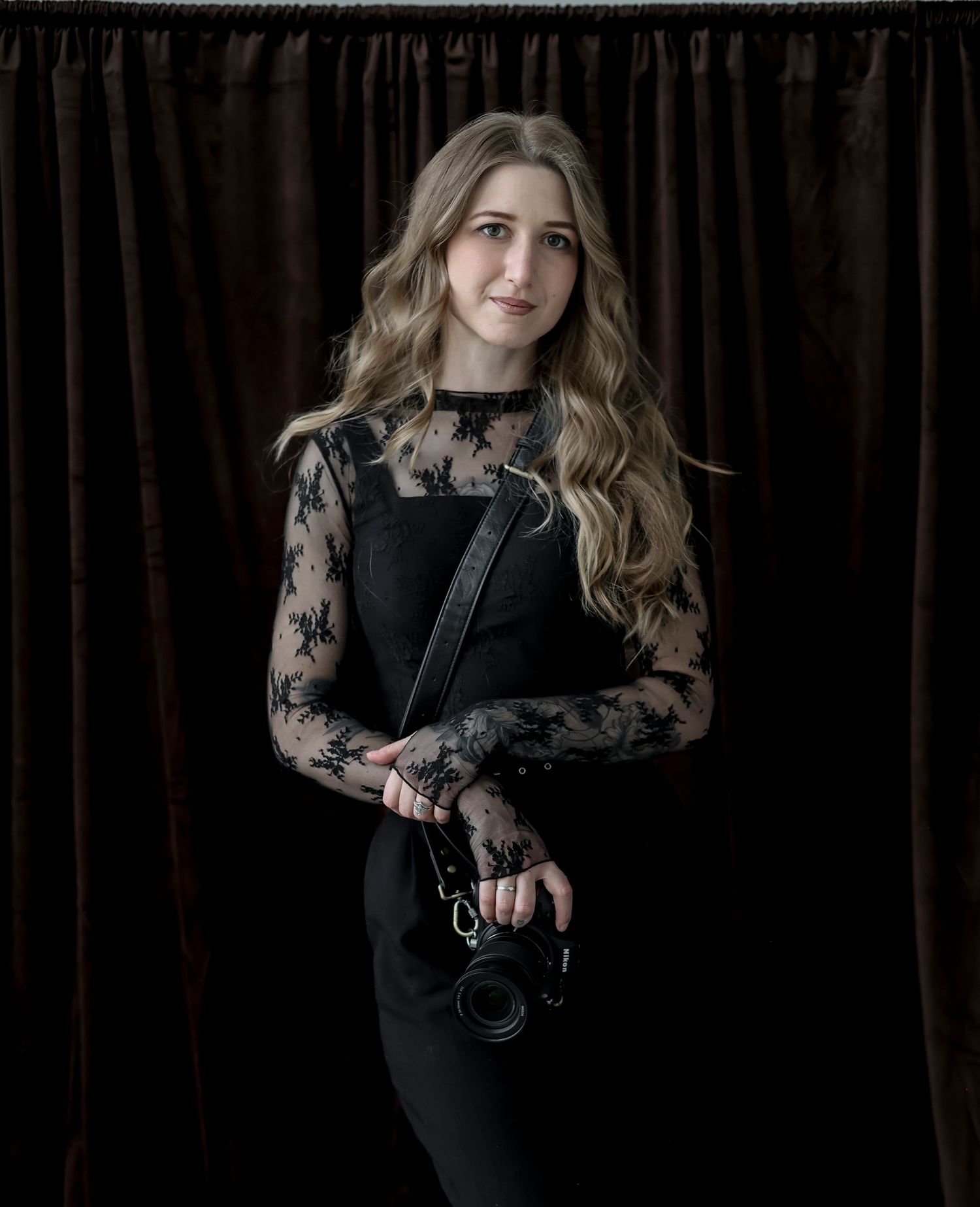 Young woman with wavy blonde hair holding a camera, wearing black lace sleeves and dark outfit against velvet curtain.