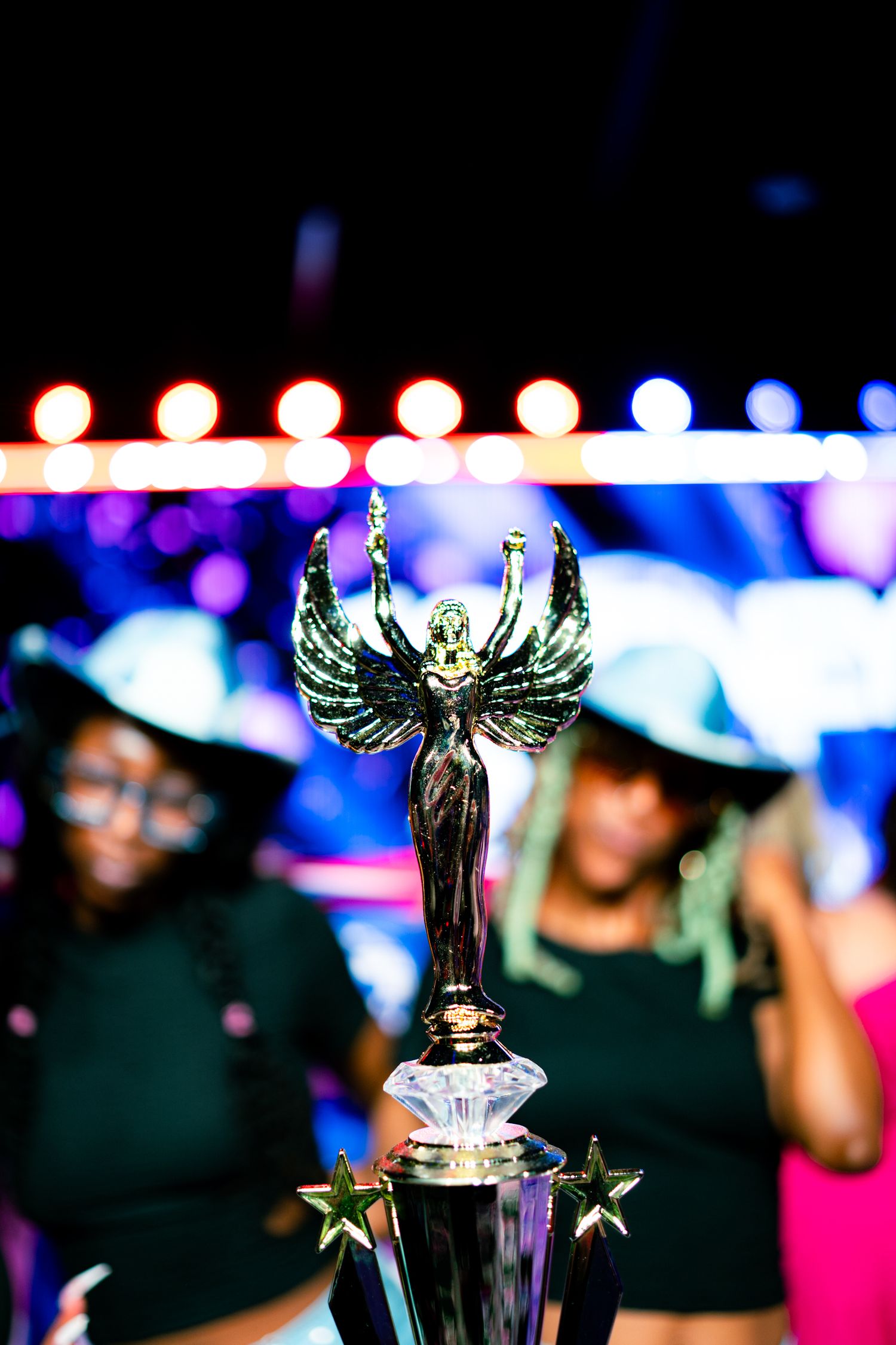 Close-up of a winged angel trophy at a dance competition with colorful stage lights in background.