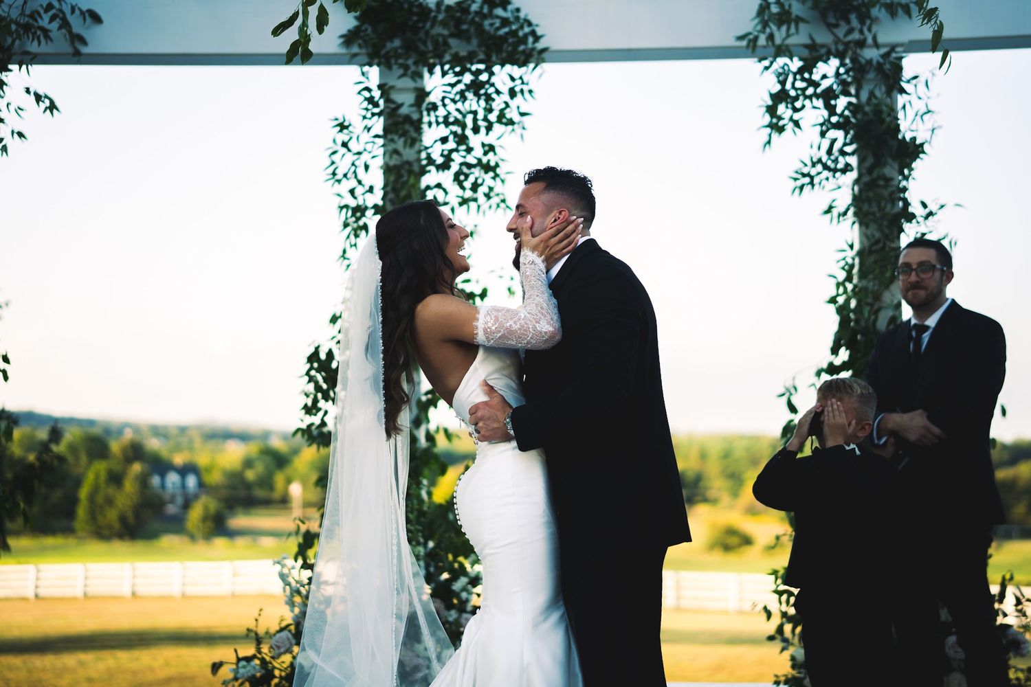 Bride and groom share first kiss under floral arch at outdoor wedding ceremony at golden hour.