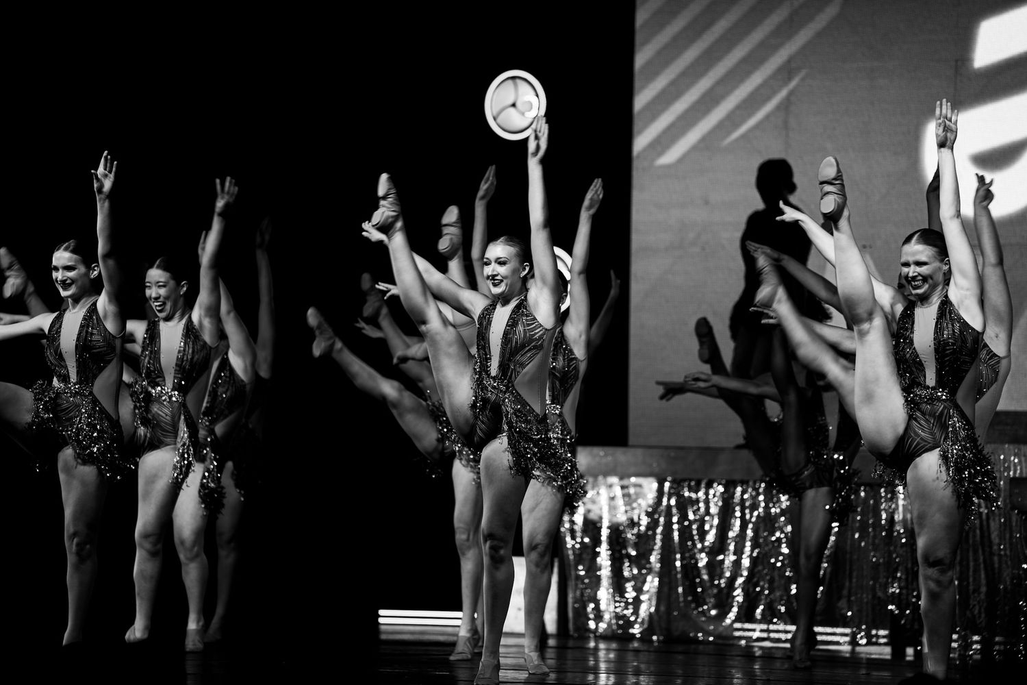 Black and white photo of dancers in sequined costumes performing on stage, one holding a trophy aloft.