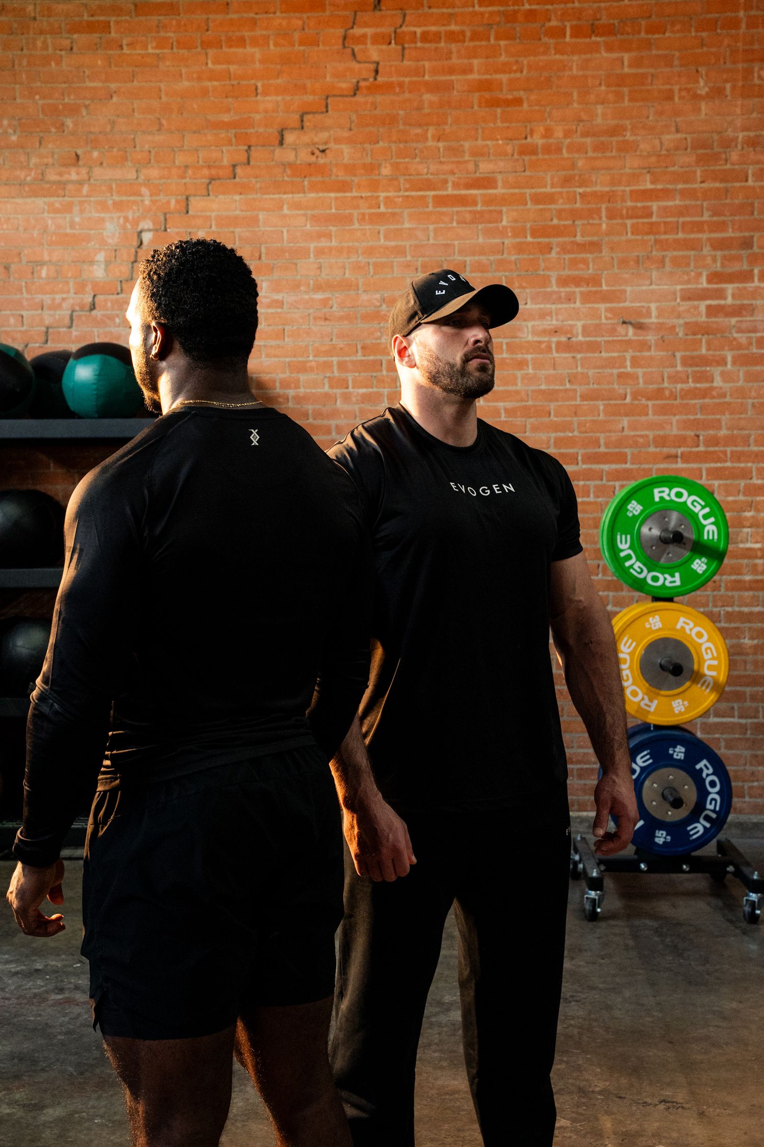 Two men in black Evogen shirts standing near colorful Rogue weight plates.
