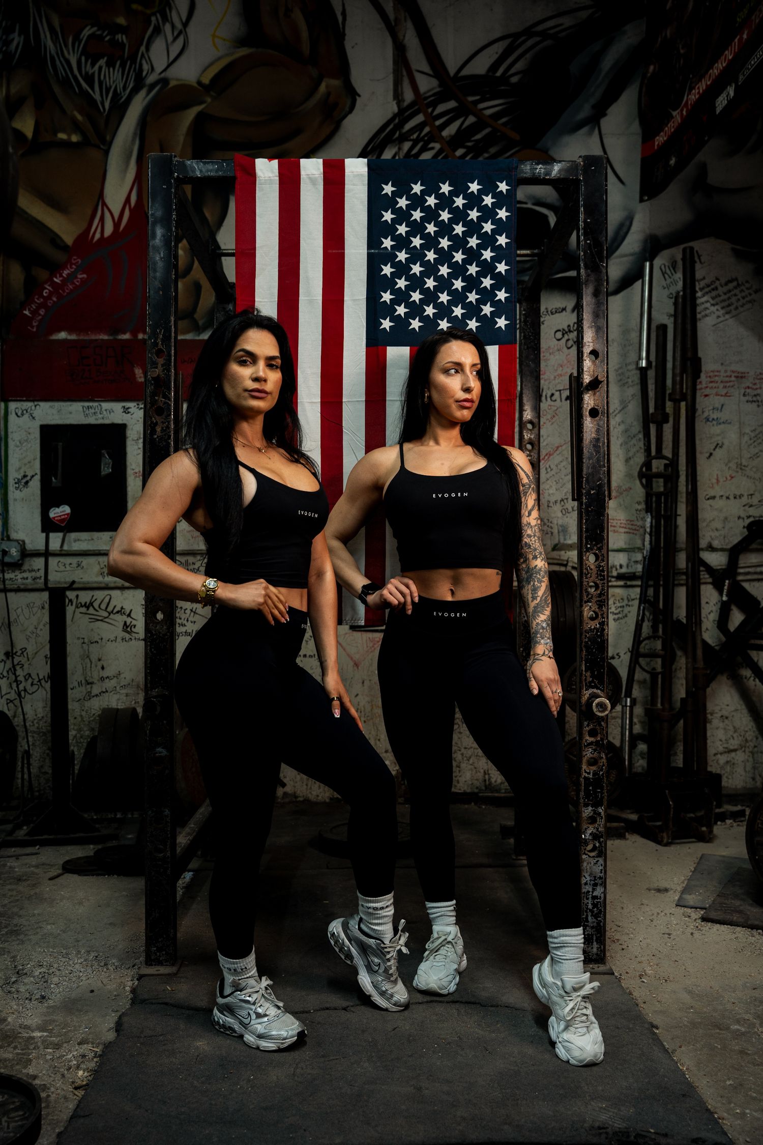 Two athletic women in black fitness wear pose in front of an American flag in a gritty gym setting.