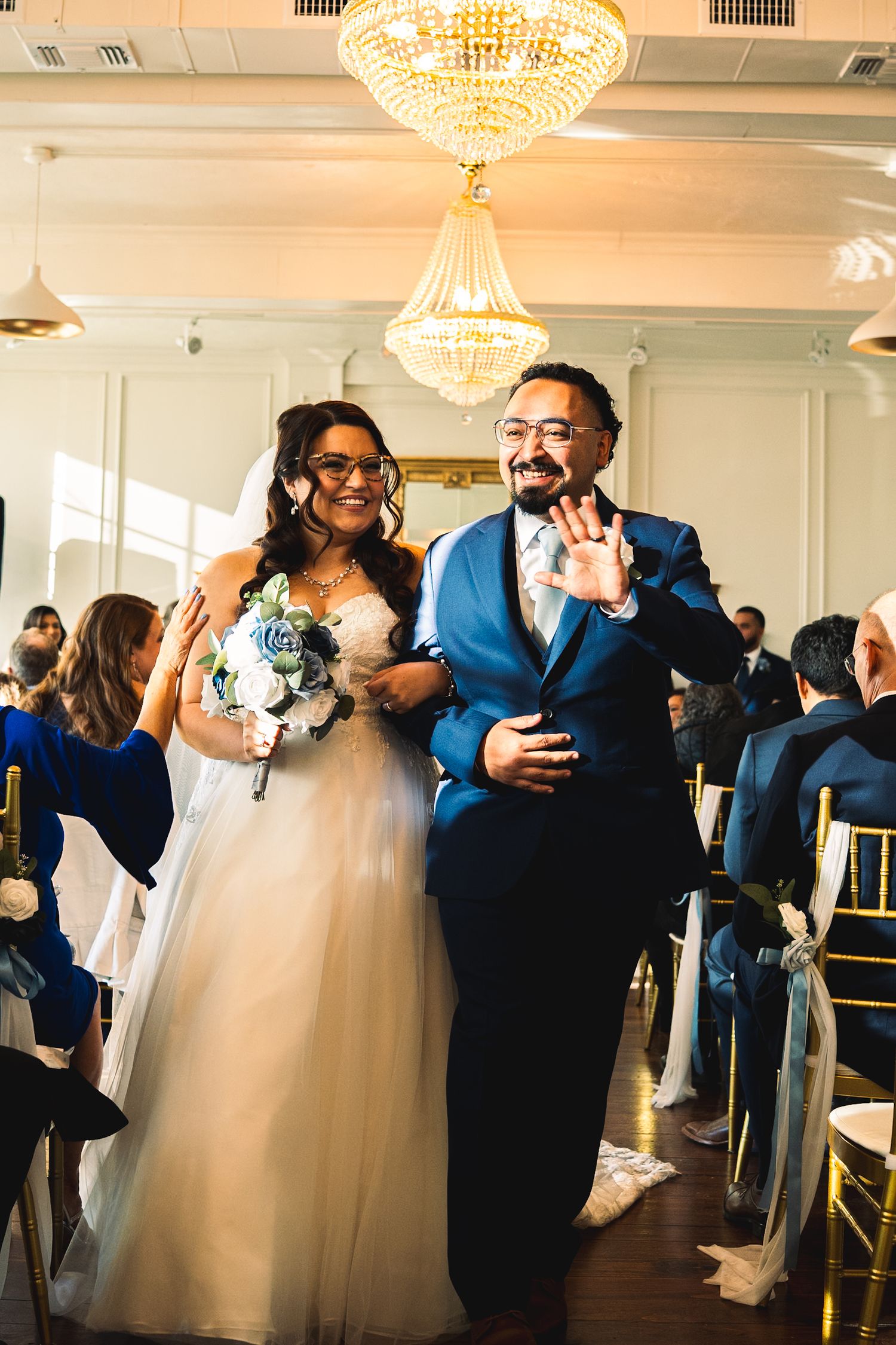 Bride and groom in glasses walk down the aisle, groom waving, holding blue bouquet.