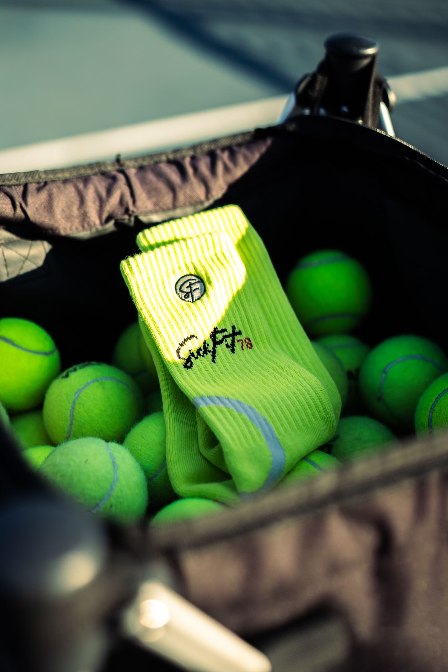Neon yellow tennis socks resting on green tennis balls in a ball hopper basket on a court.