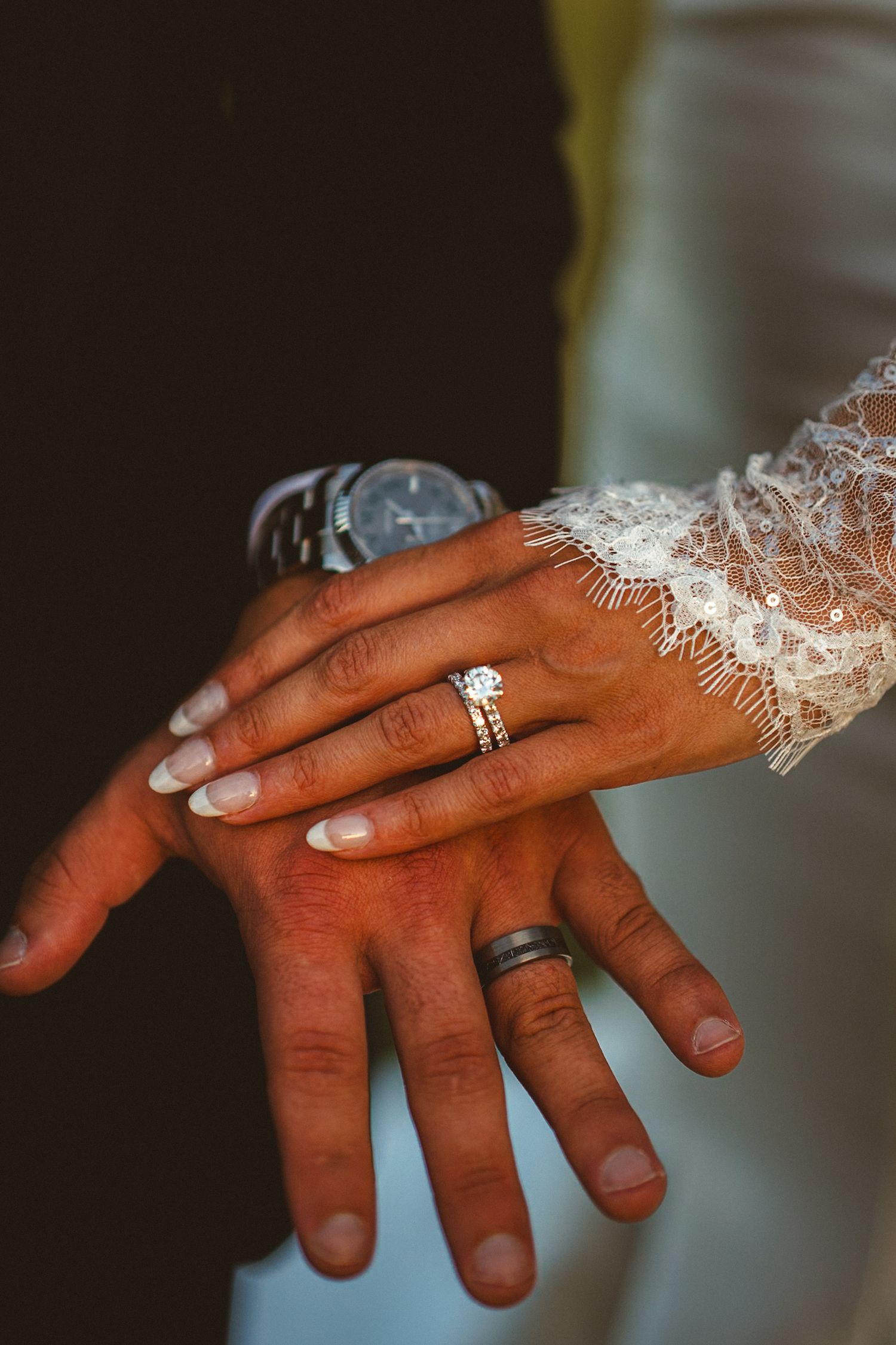 Close-up of newlyweds hands showing diamond engagement ring, wedding bands, lace bridal sleeve and grooms watch.