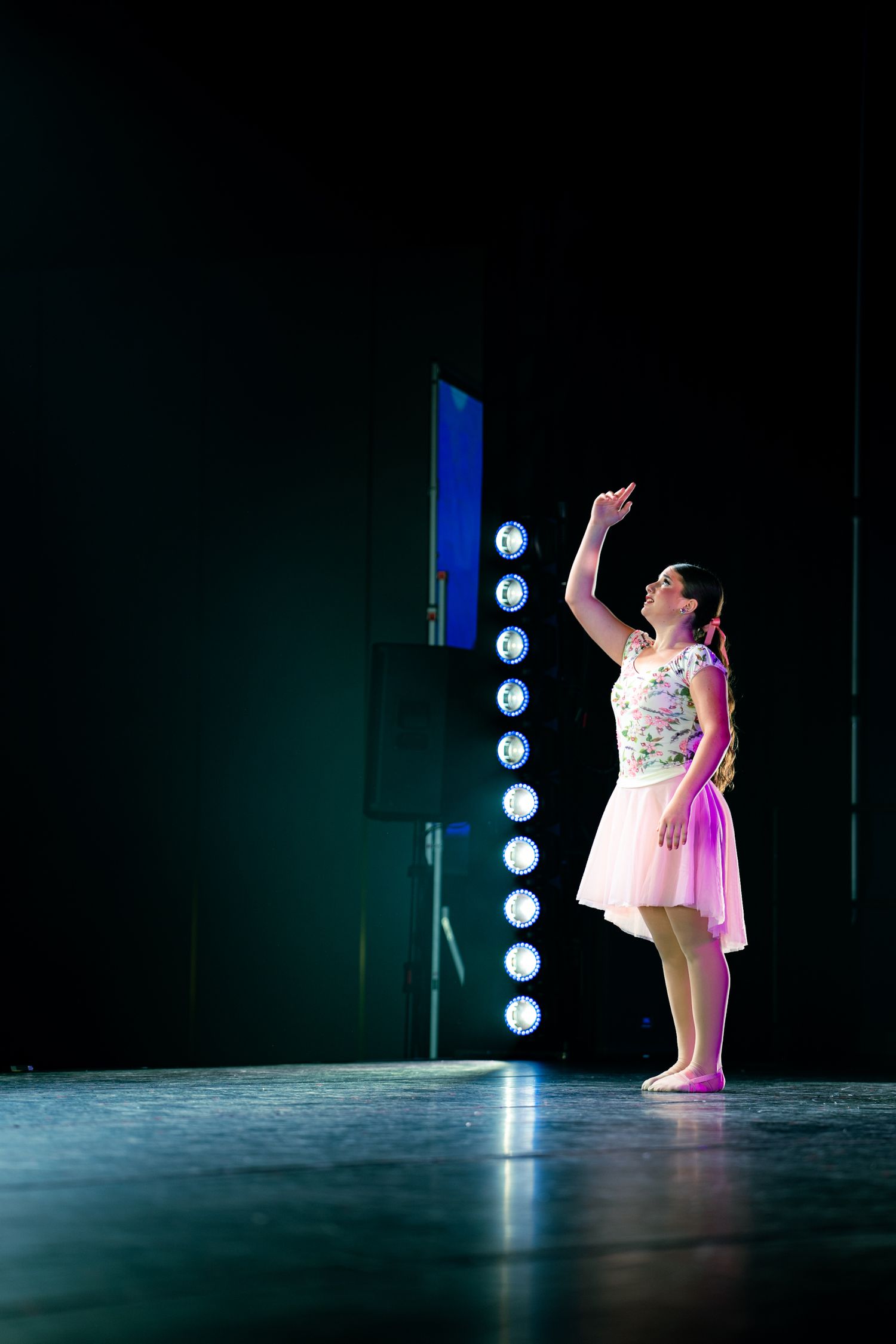 Young girl in pink skirt performs on stage with raised arm, illuminated by blue stage lights in dark theater.