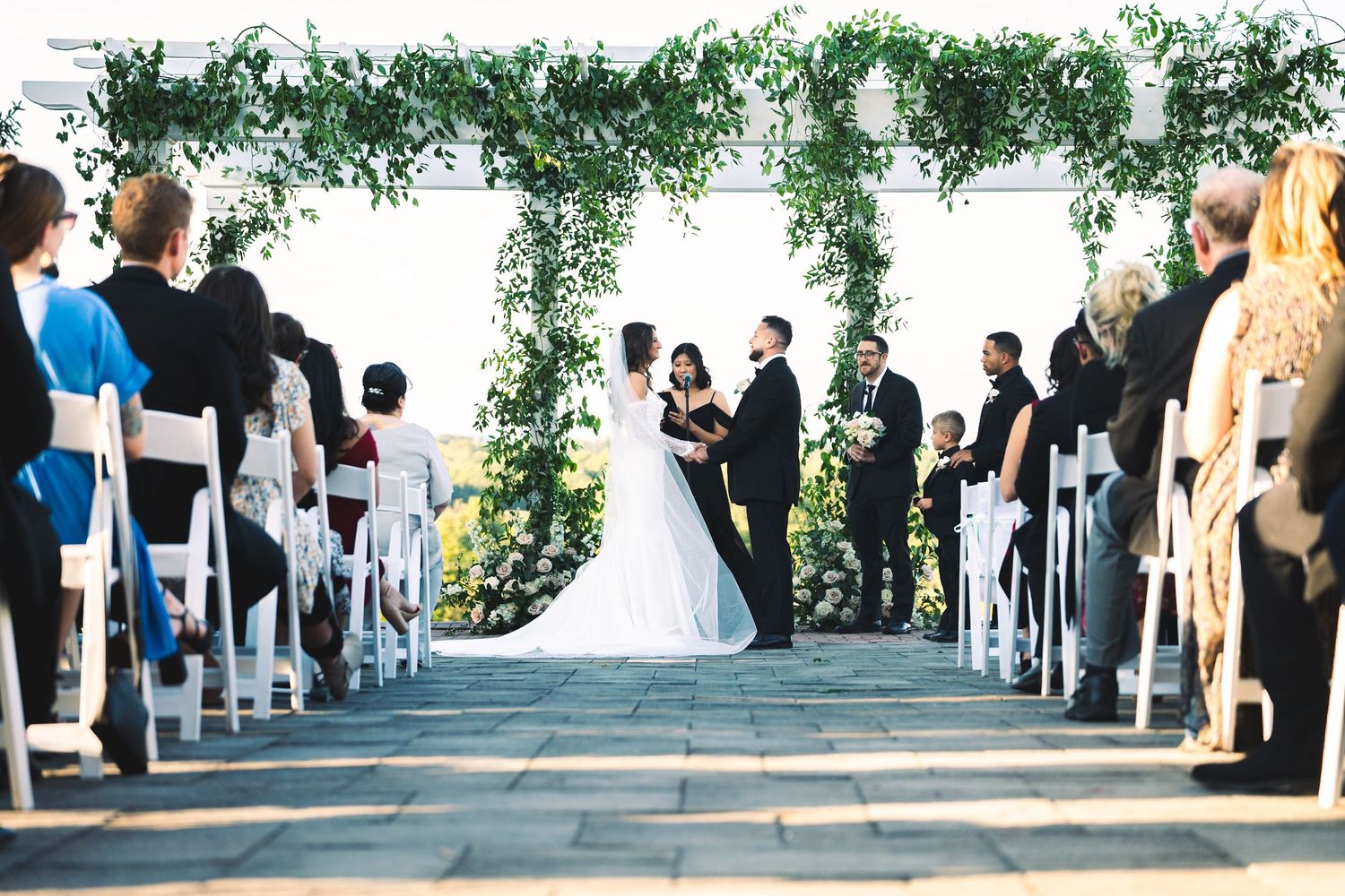 Bride and groom exchange vows under lush greenery arch at outdoor wedding ceremony with white chairs.