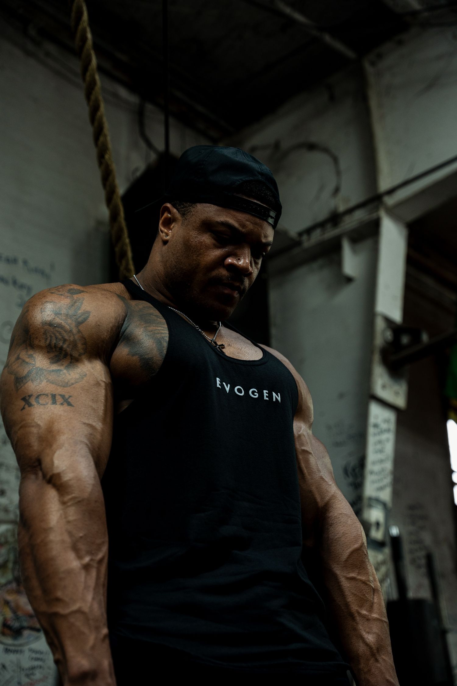 A heavily tattooed muscular man in a black Evogen tank top poses in a gritty gym setting.